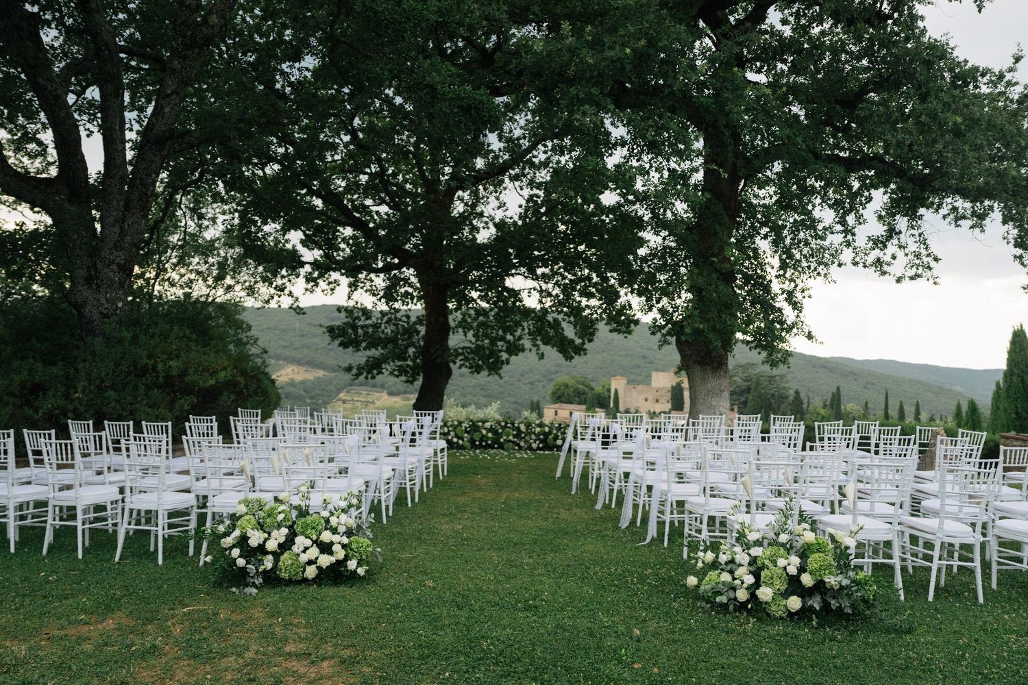 Elegant outdoor wedding ceremony setup with white Chiavari chairs and floral arrangements under large oak trees.