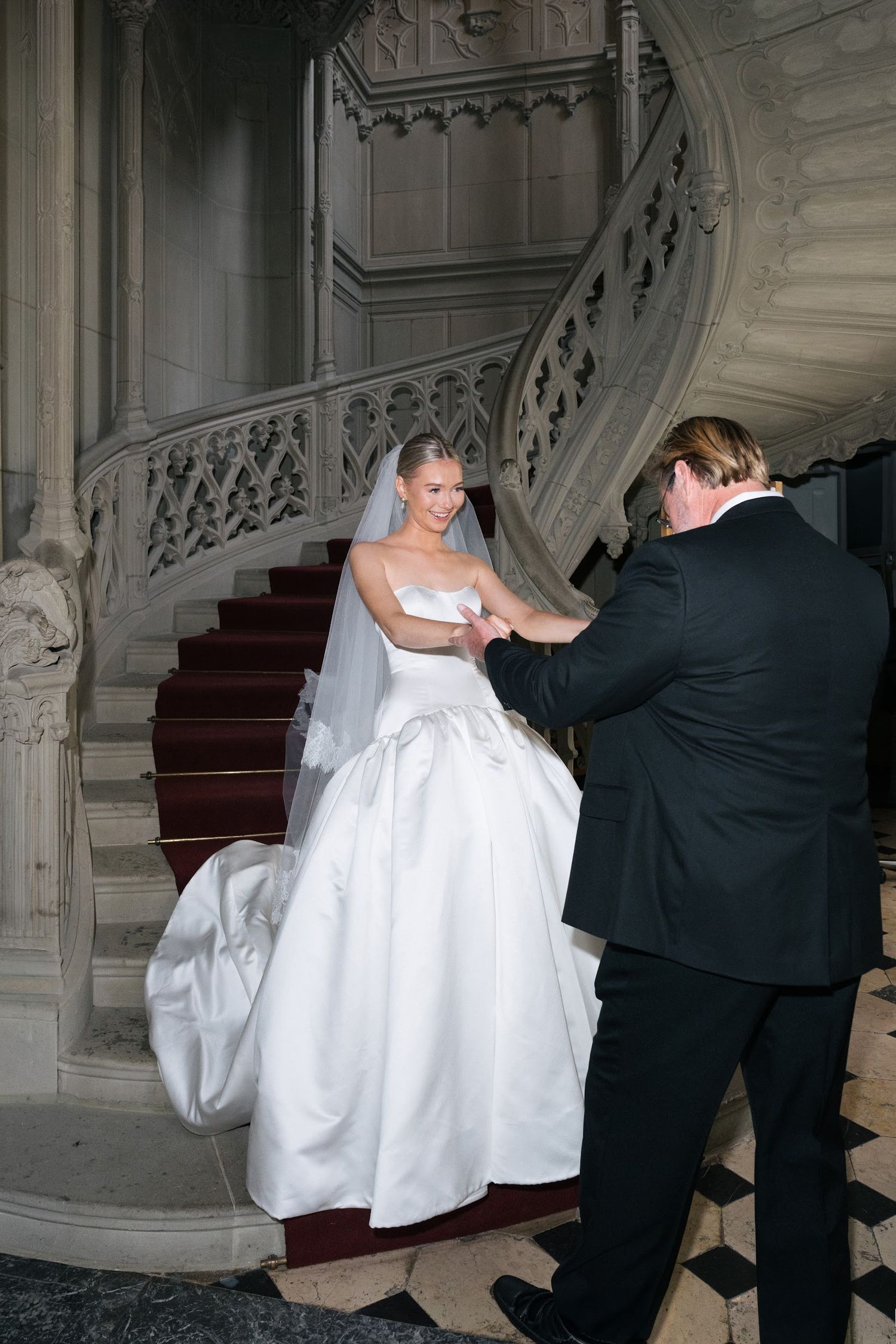 Bride in strapless white ball gown holding hands with suited gentleman on elegant marble spiral staircase.