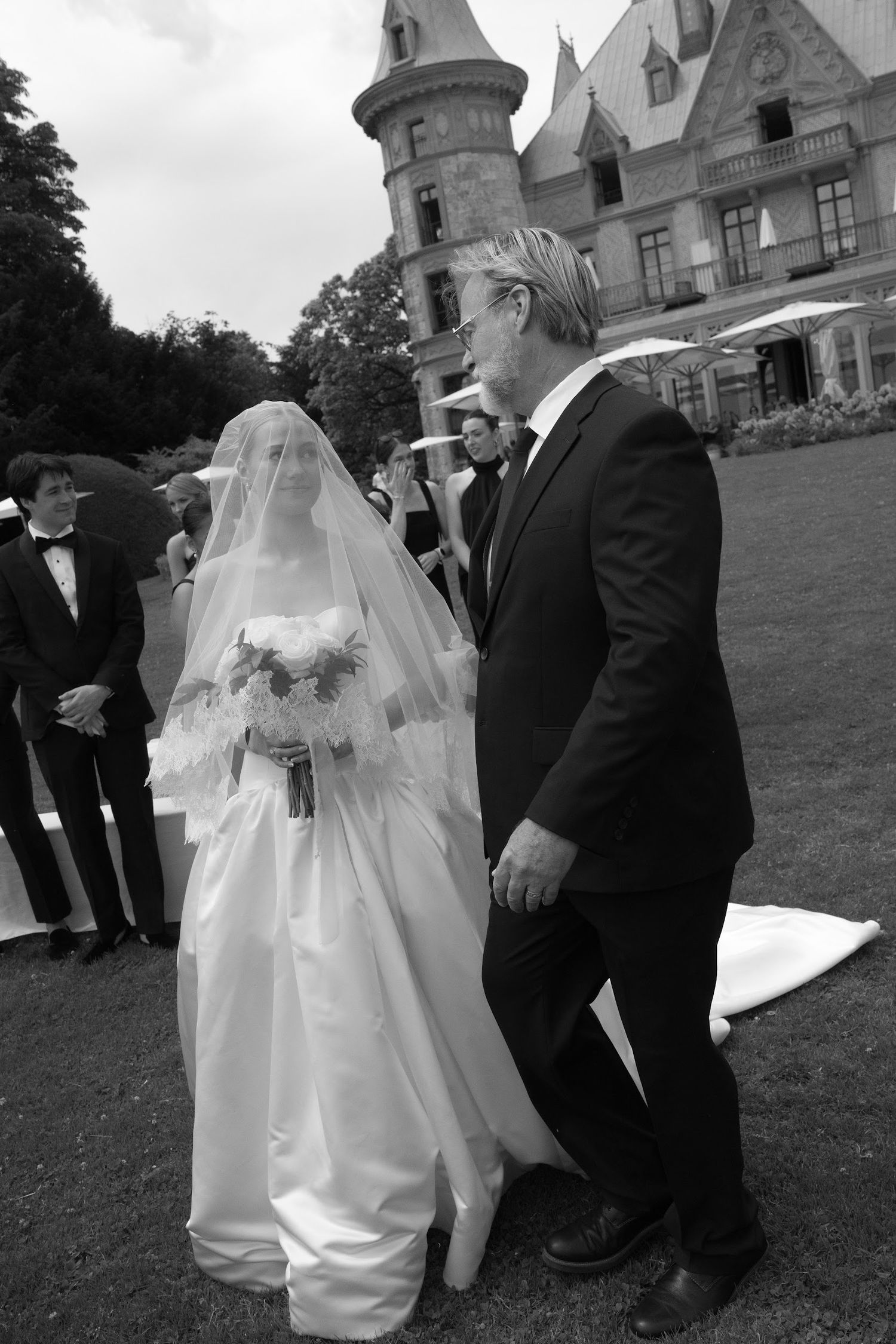 Black and white photo of a bride in a flowing gown and veil walking with her father toward the wedding ceremony.