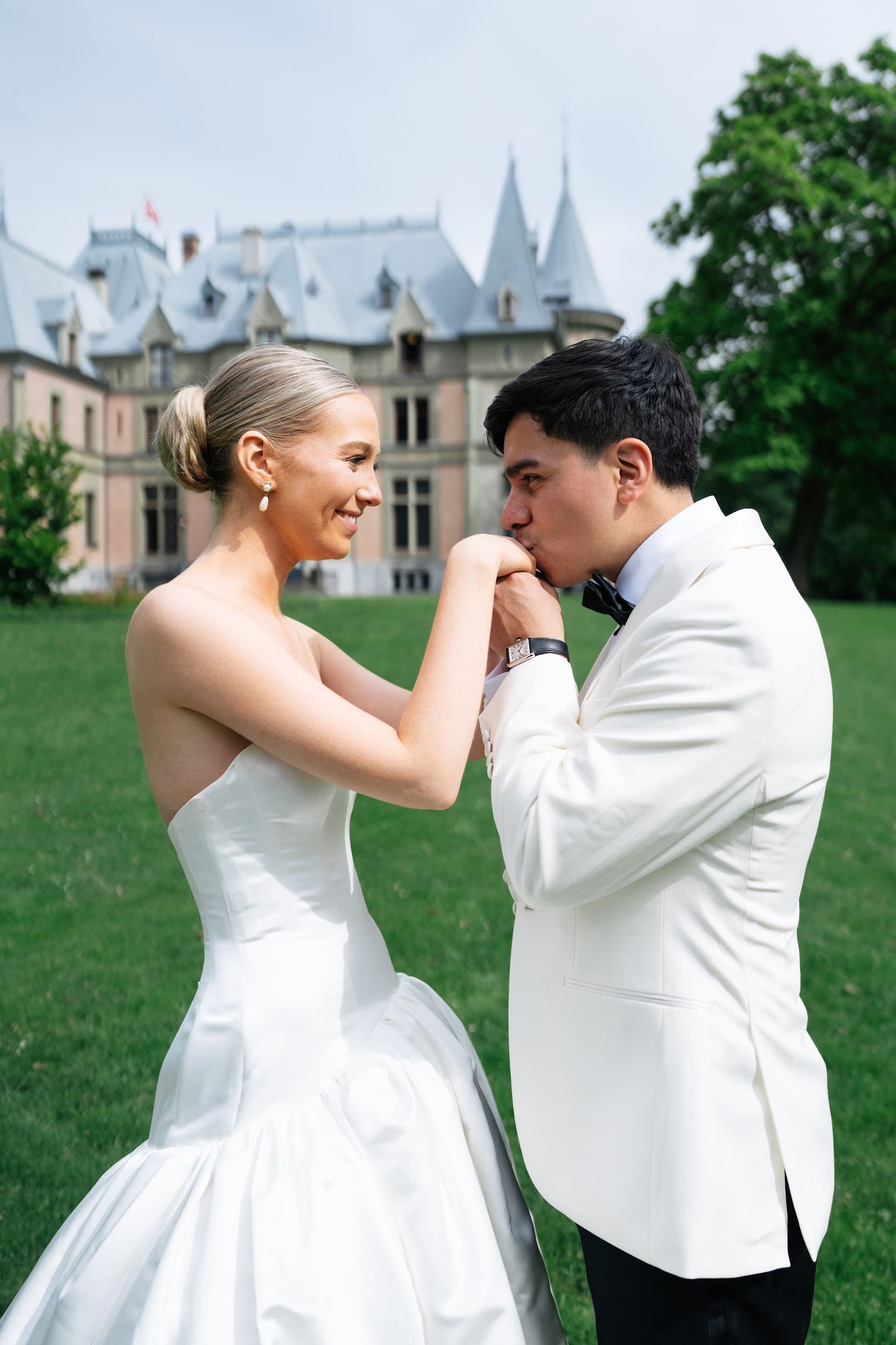 Groom in white tuxedo kissing bride's hand outside a French château on their wedding day.