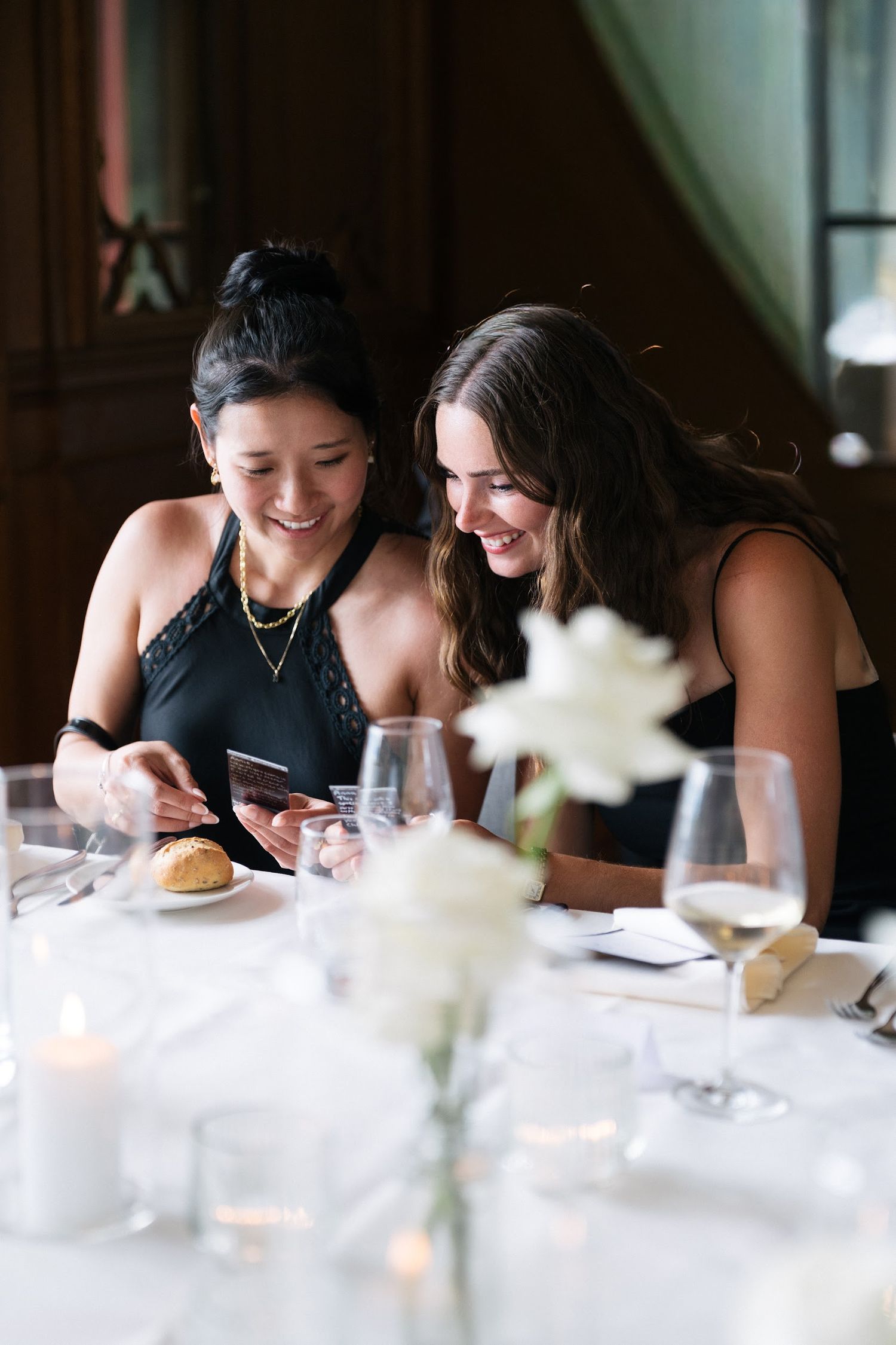 Two women in black dresses smile while looking at a phone during an elegant wedding reception dinner.
