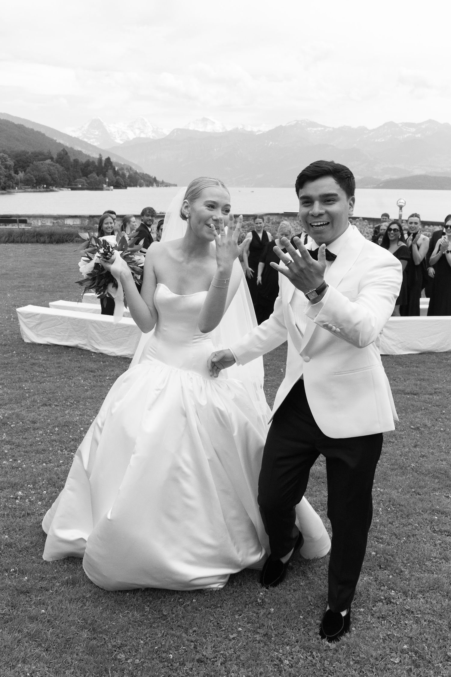 Black and white photo of newlyweds joyfully showing their wedding rings to guests after their outdoor ceremony.