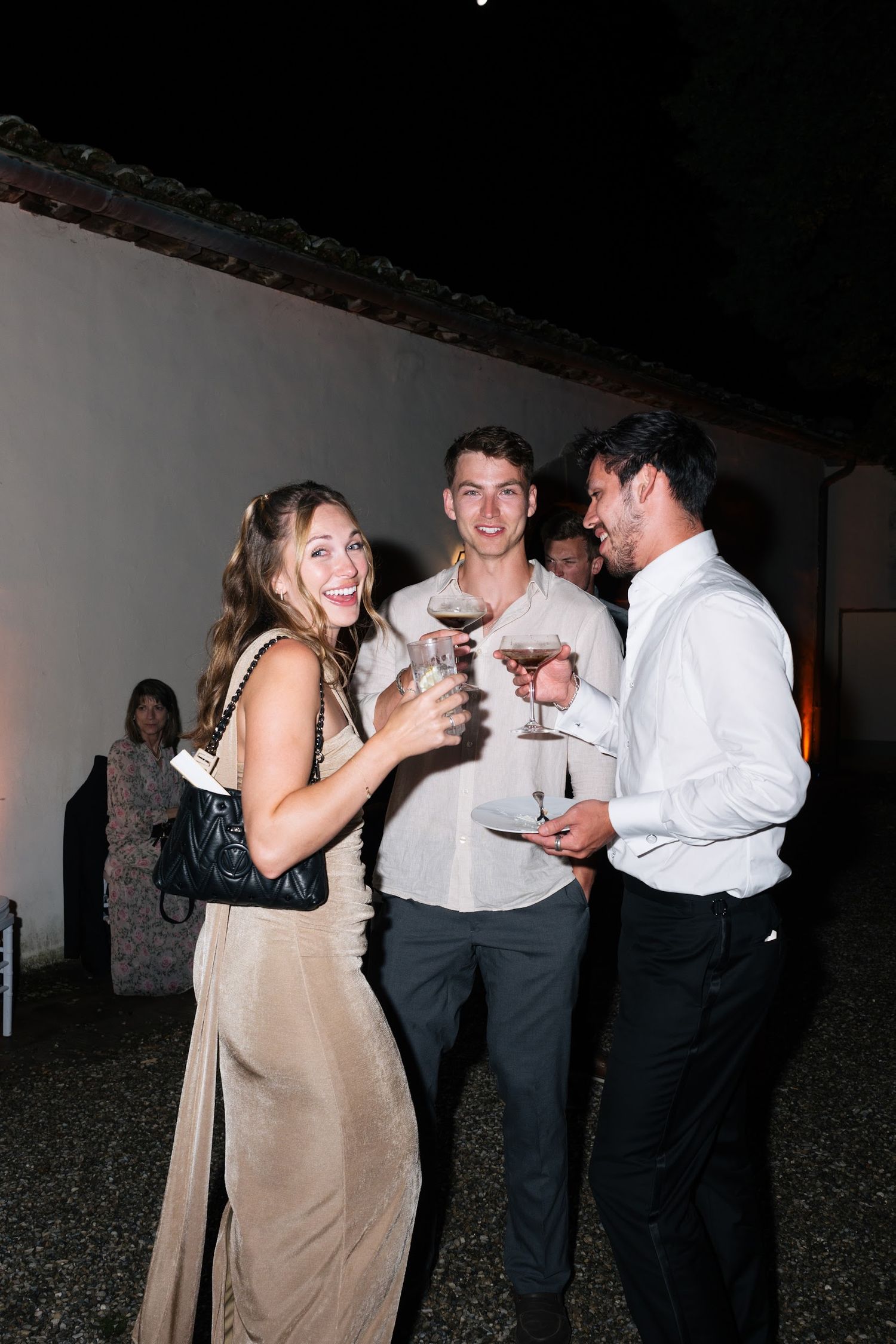 Three wedding guests toasting with wine glasses and smiling at an outdoor evening reception.