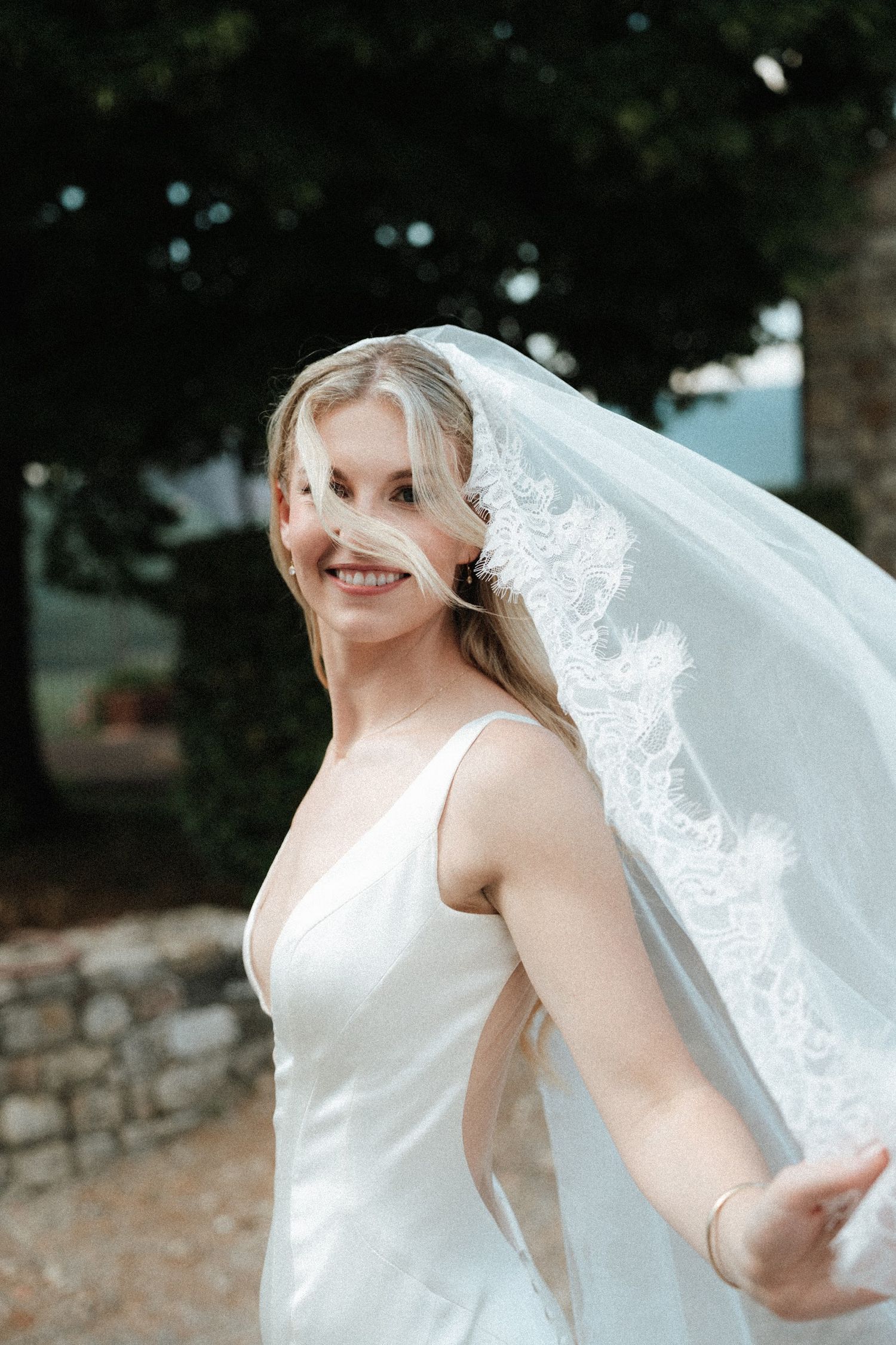 Smiling bride twirls in white gown with lace veil blowing across her face in outdoor setting.