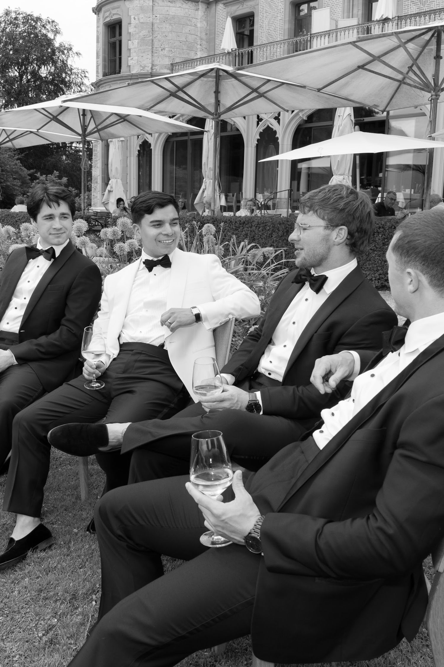 Black and white photo of groom in white tuxedo jacket laughing with groomsmen in black tuxedos at outdoor wedding reception.