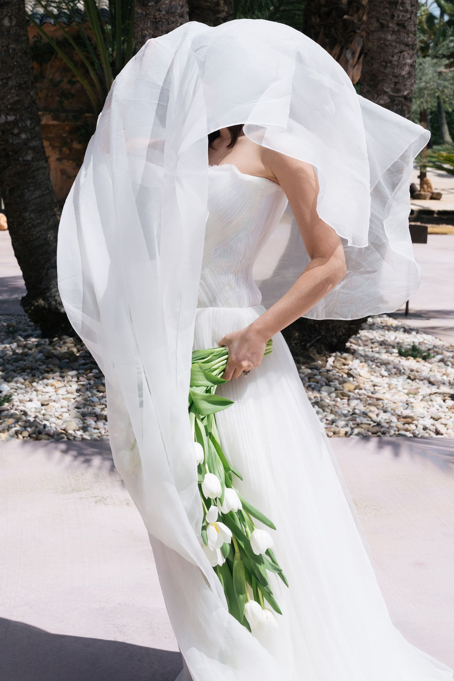Bride in white gown holds tulip bouquet as her veil billows dramatically in the breeze outdoors.