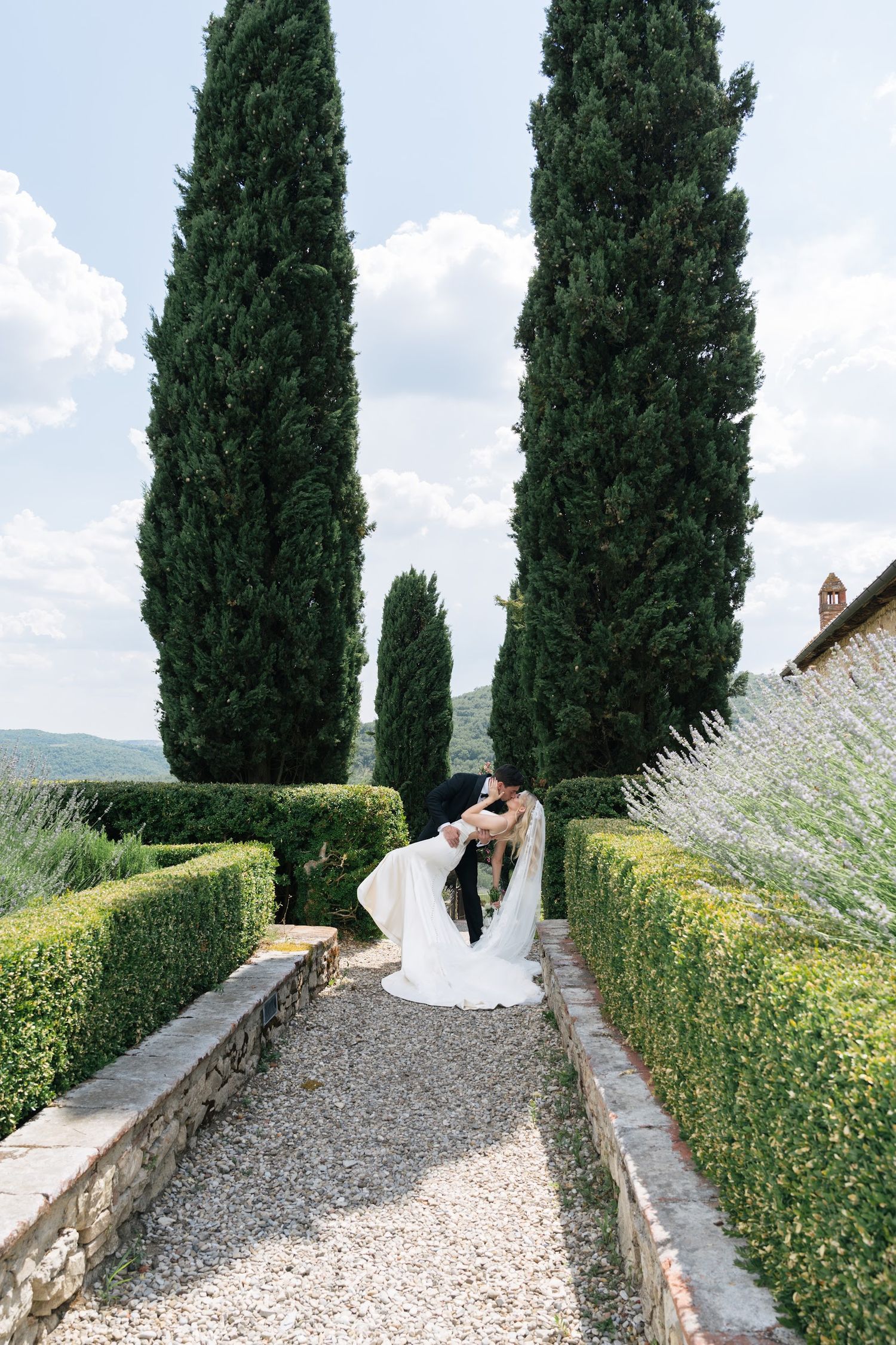 A bride and groom share a romantic dip kiss in a formal Italian garden with tall cypress trees.