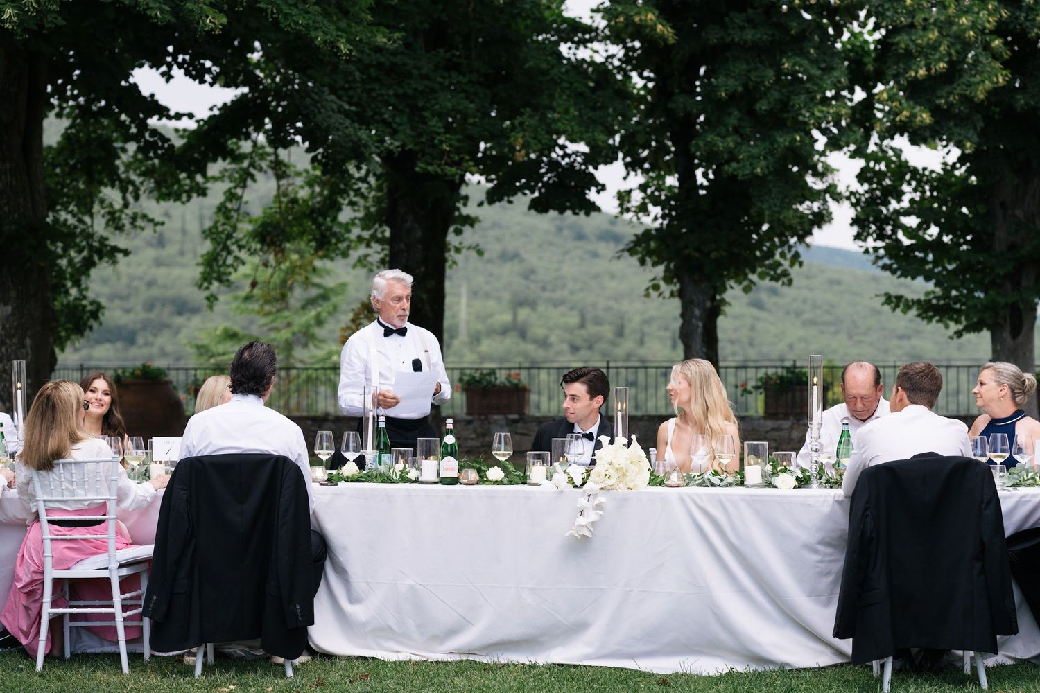Wedding speaker addresses guests at a beautifully set outdoor Tuscan reception table under green trees.