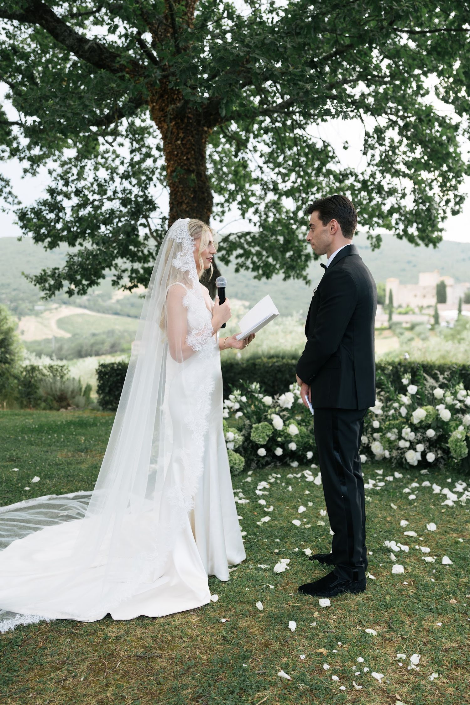 Bride reads wedding vows to groom beneath oak tree surrounded by white floral arrangements in Tuscany.