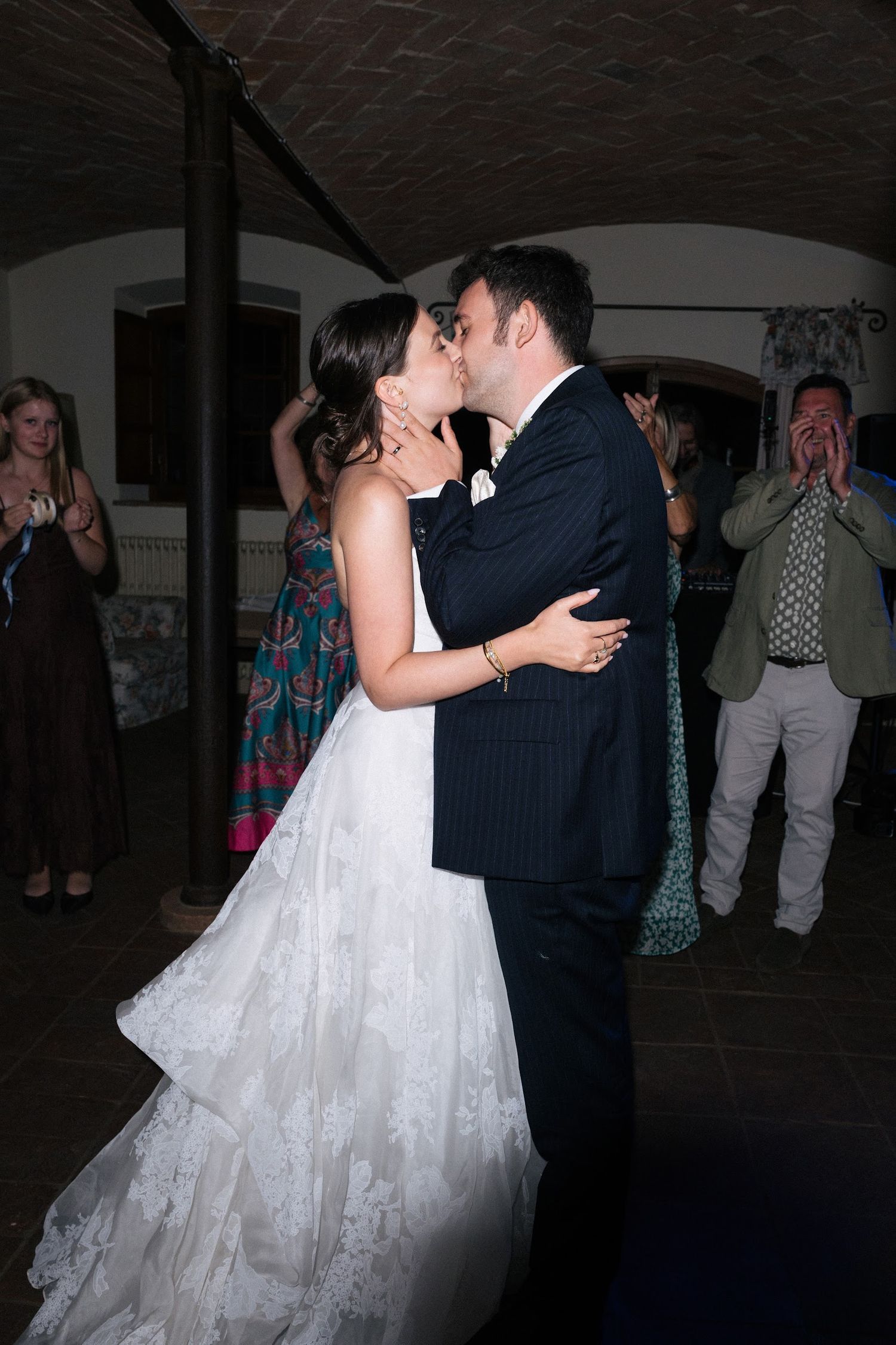 Bride and groom share first kiss on wedding dance floor surrounded by guests.