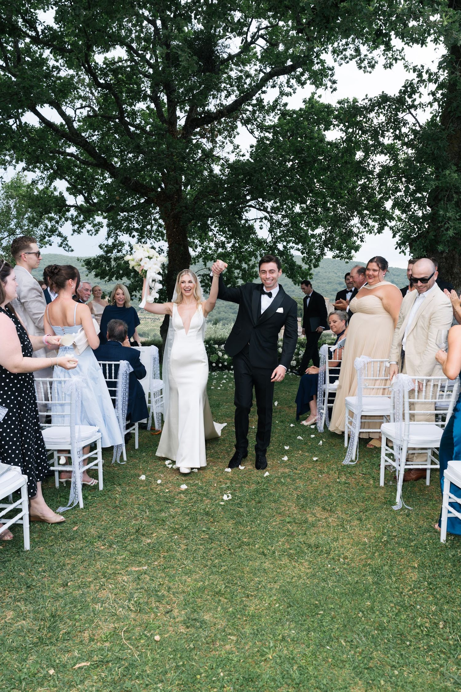 Newlyweds celebrate recessional with raised hands amid cheering guests at outdoor Tuscan wedding ceremony.