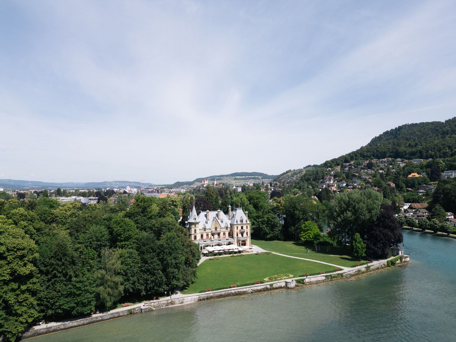 Aerial view of an elegant lakeside castle surrounded by lush green trees on the shores of a Swiss lake.