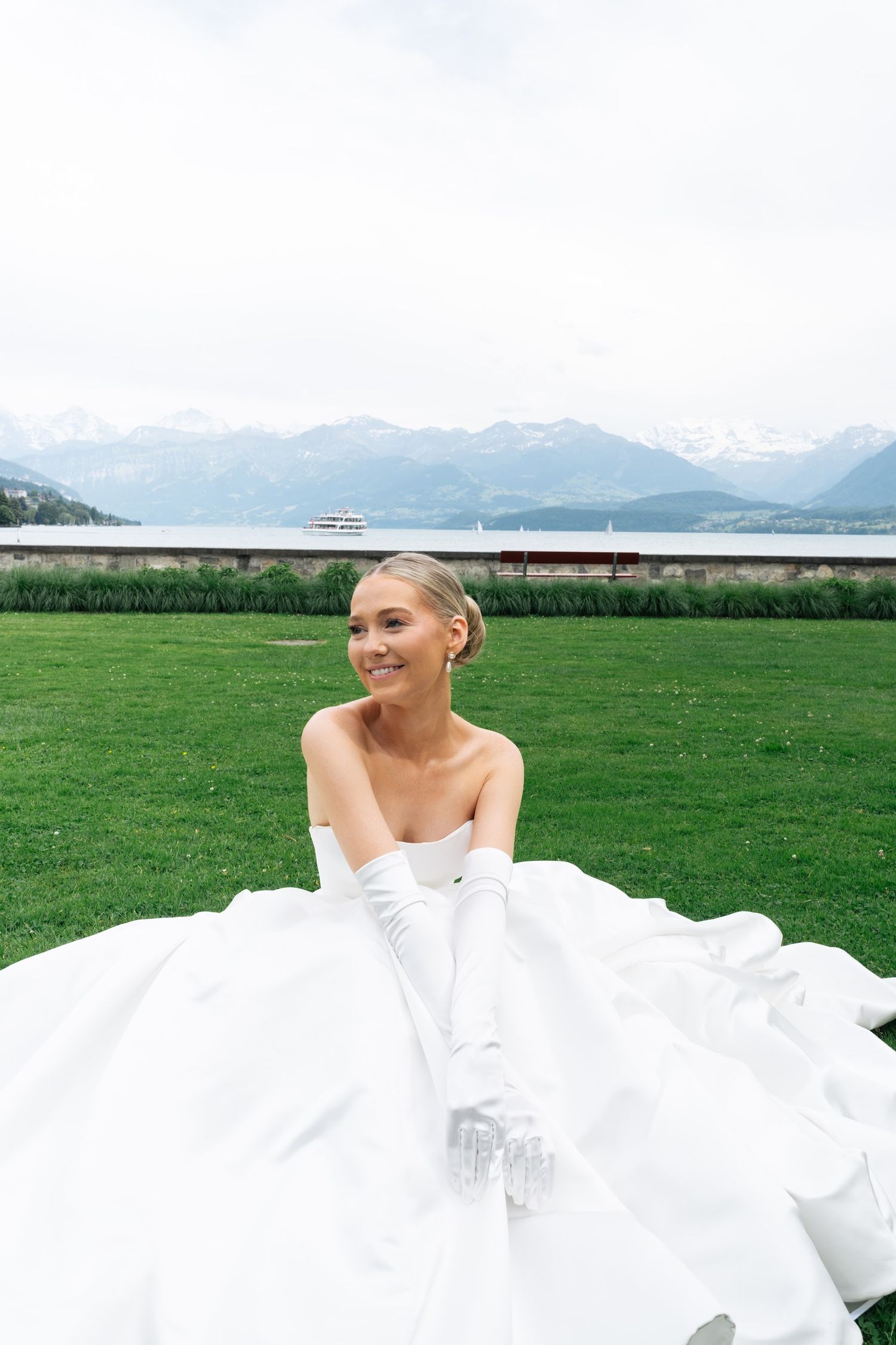 Smiling bride in strapless white gown and long gloves sitting on grass with alpine lake backdrop.