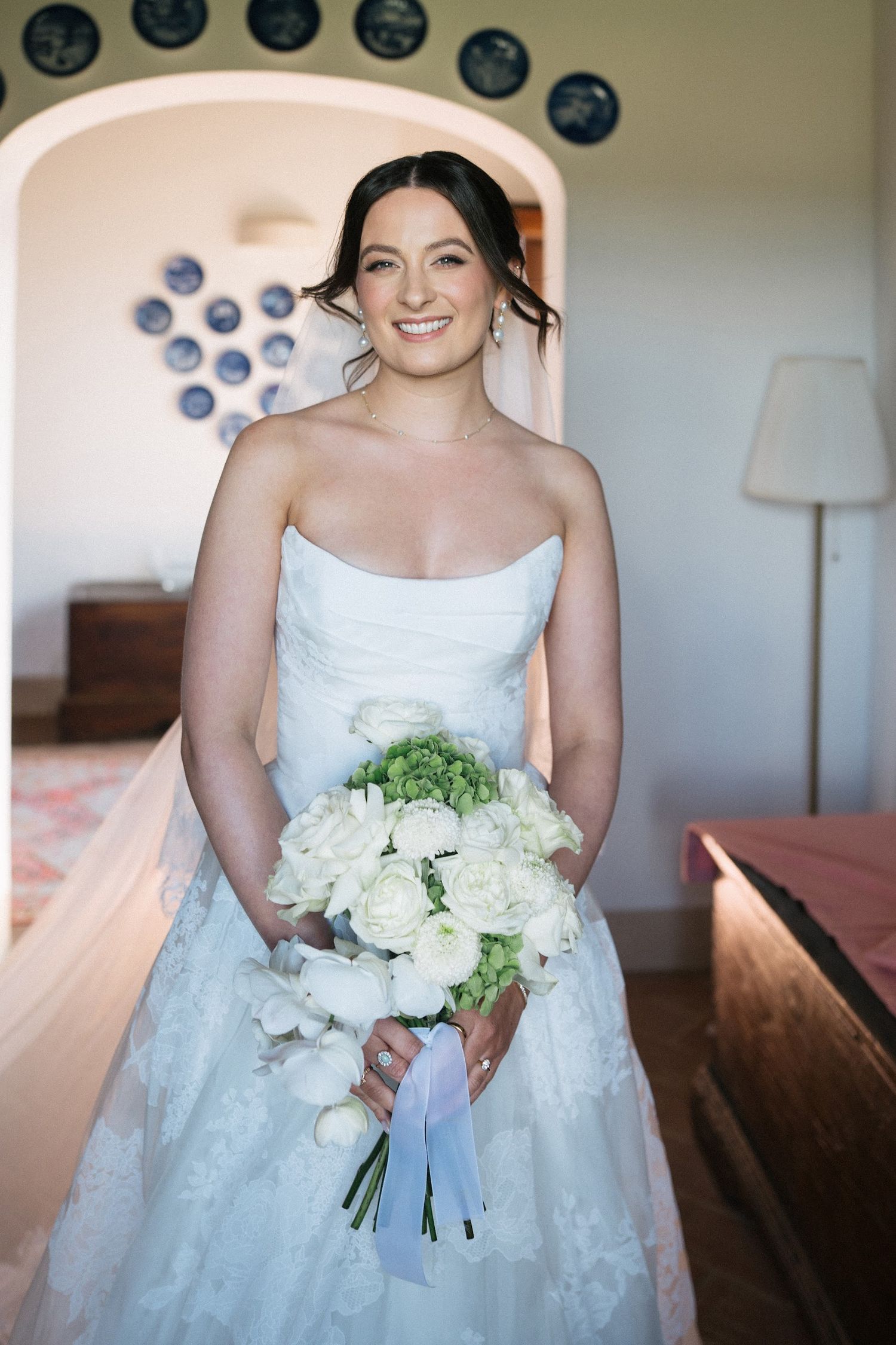 Smiling bride in strapless lace wedding dress holding white and green bouquet in elegant Italian villa.