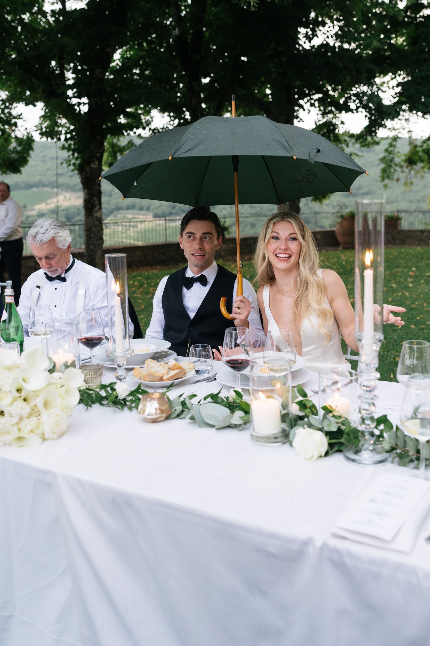 Smiling bride and groom sitting at wedding reception table under umbrella during light rain.