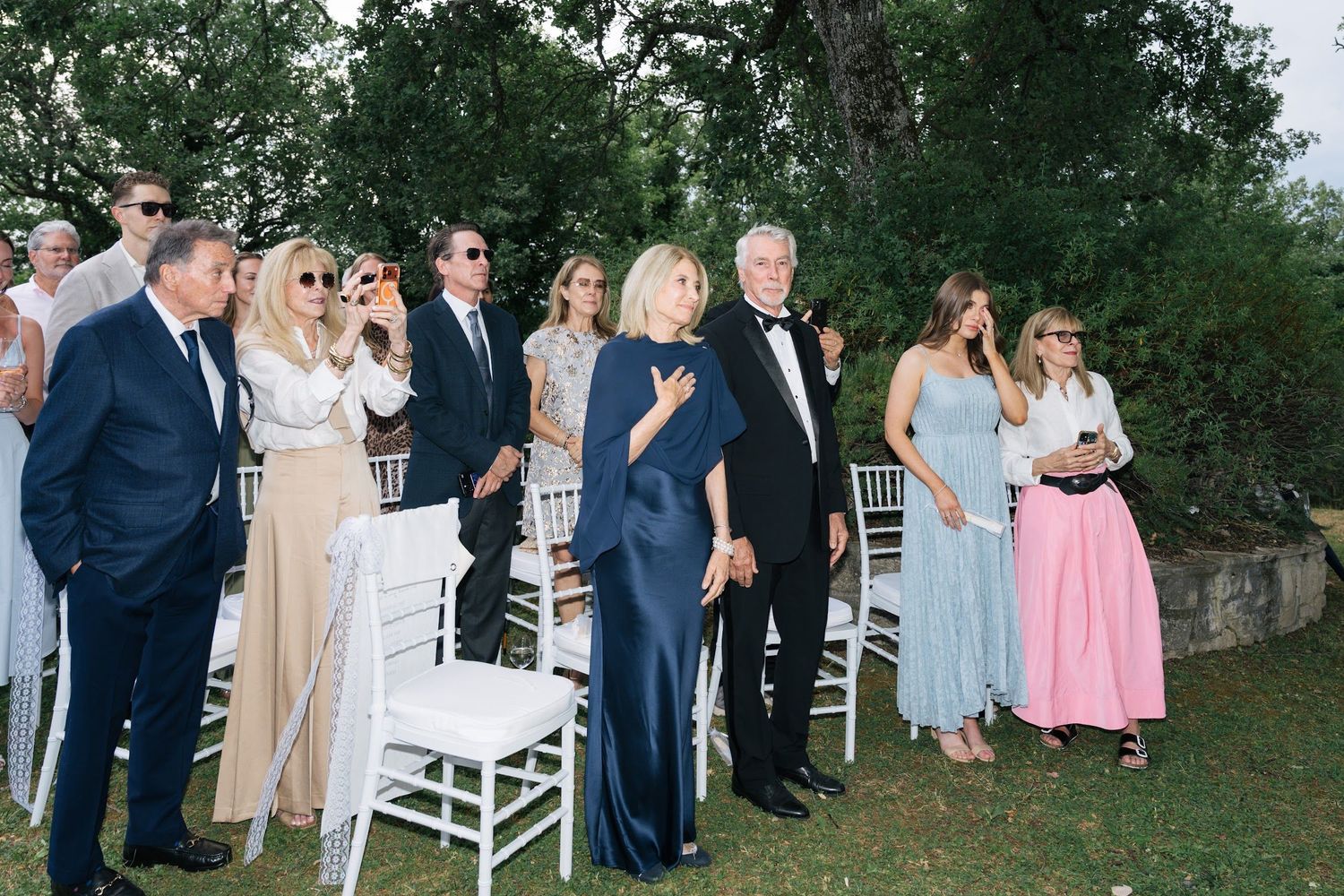 Wedding guests in elegant formal attire stand and watch the outdoor ceremony at a Tuscan villa wedding.