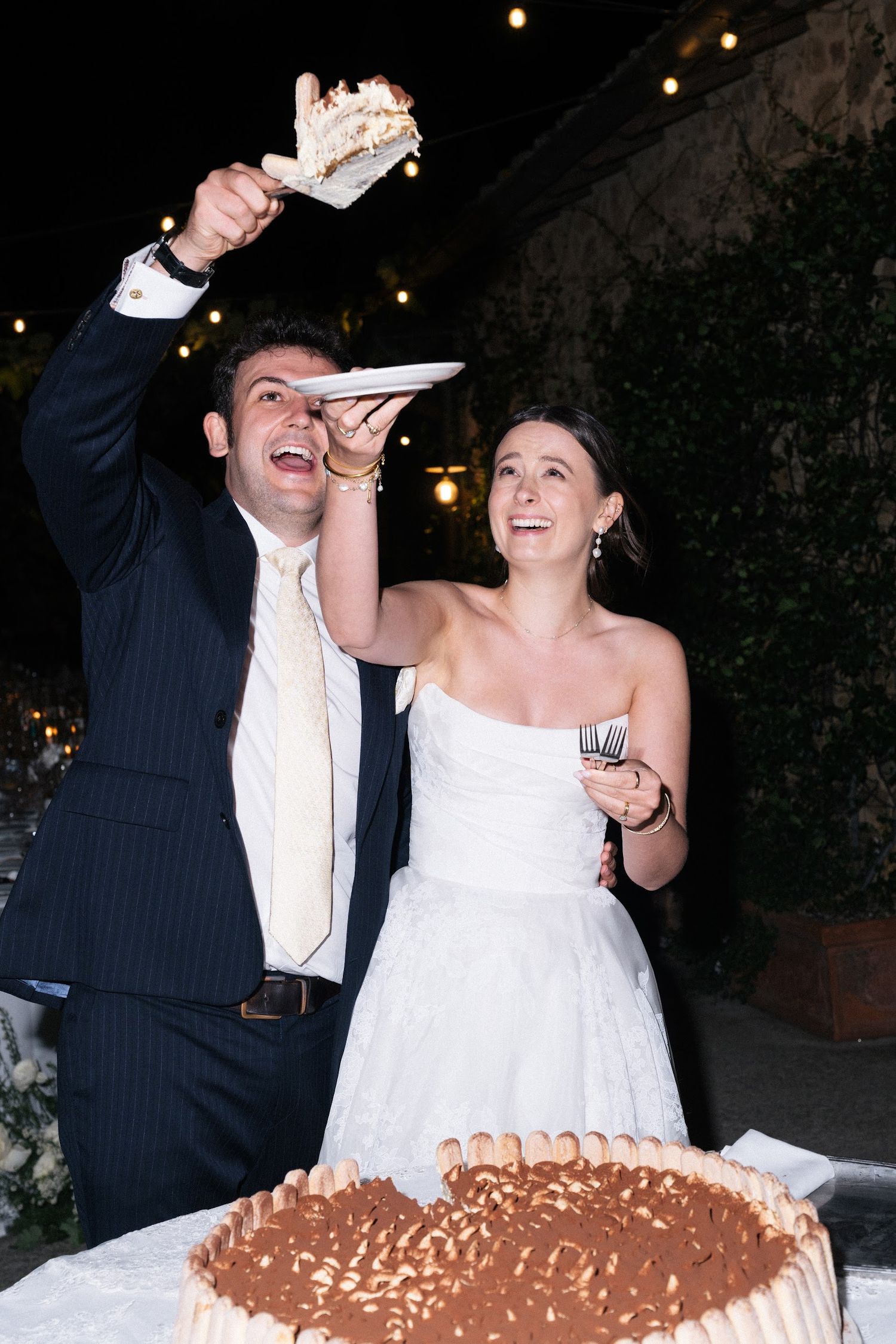 Groom proudly lifts a slice of tiramisu wedding cake beside smiling bride.