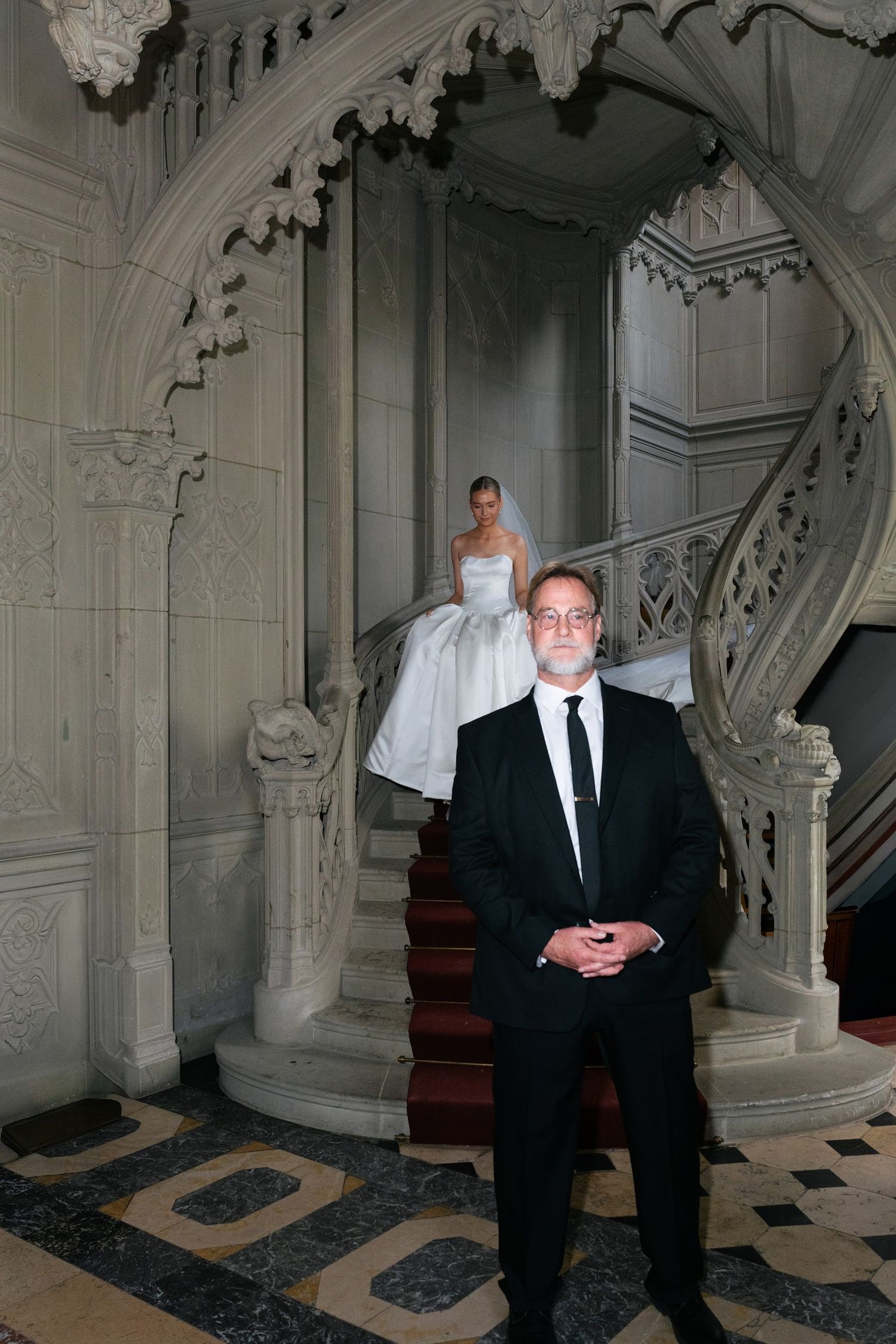 Bride descending ornate gothic spiral staircase in flowing white gown with father waiting below.