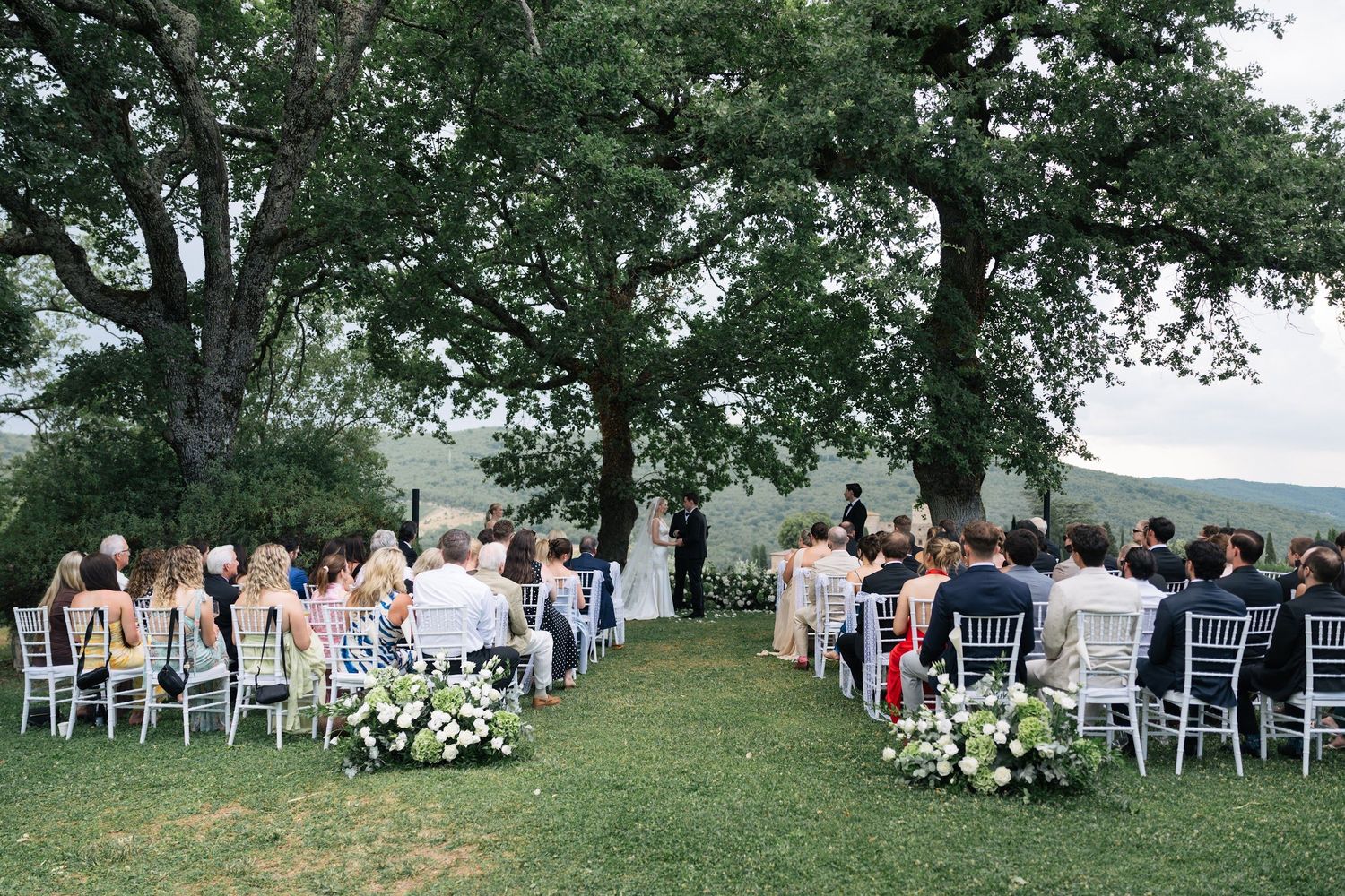 Elegant outdoor wedding ceremony under large oak trees with rolling Tuscan hills and white floral arrangements.