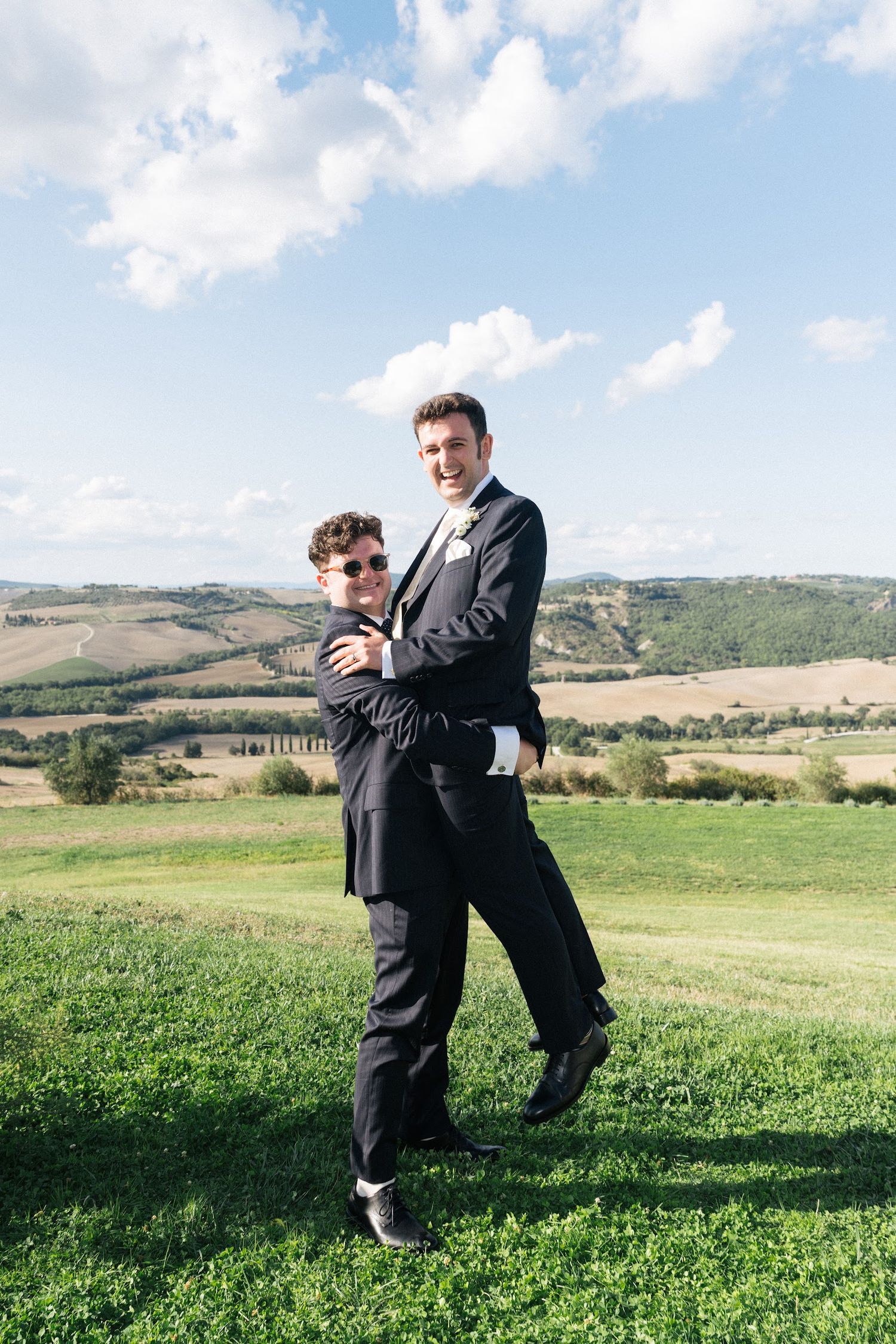Groom and groomsman playfully posing together on green hillside with scenic Tuscan valley backdrop.