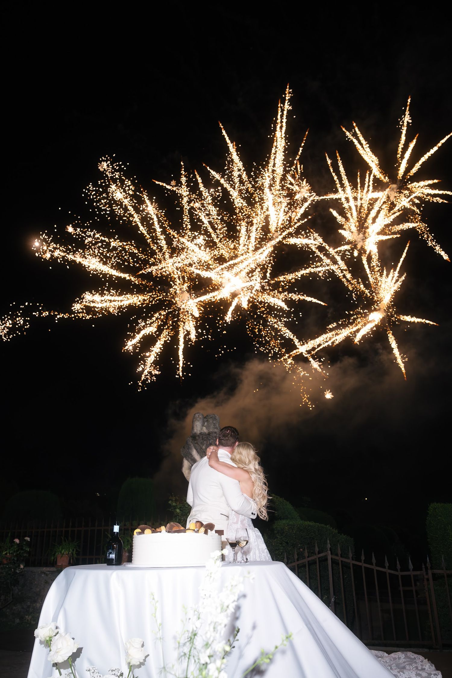 Couple kisses beside wedding cake as spectacular fireworks explode in night sky above them.