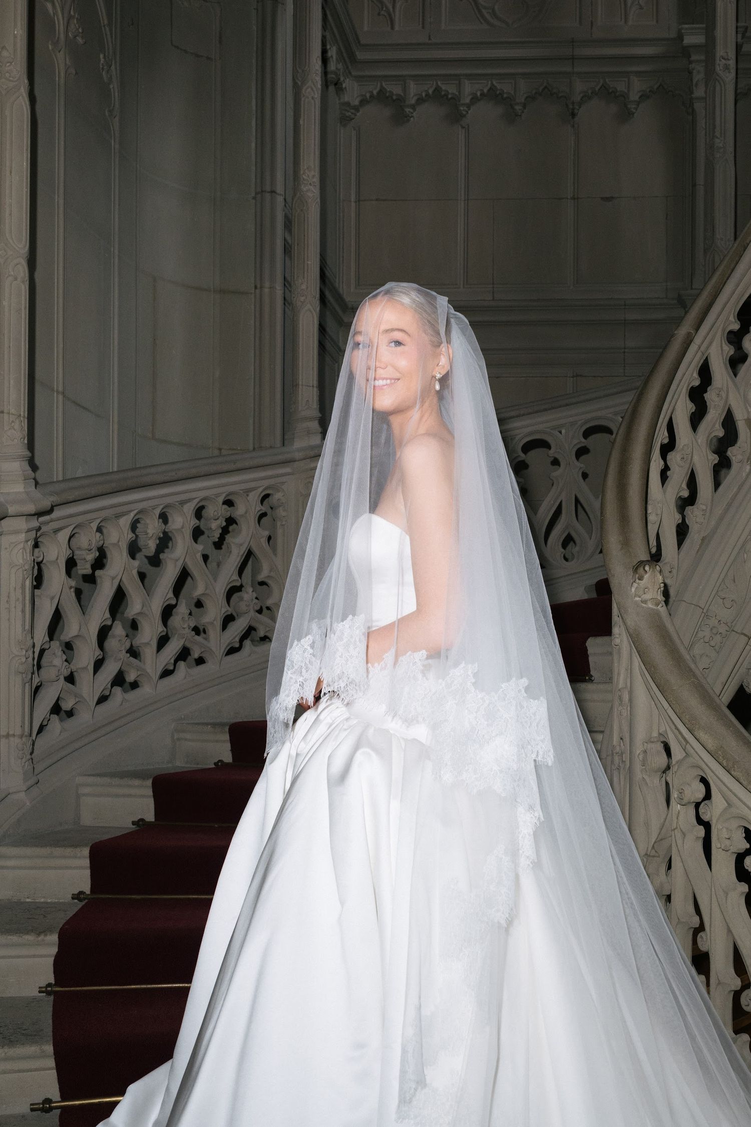 Smiling bride in strapless satin ball gown with long veil posing on ornate Gothic marble staircase.