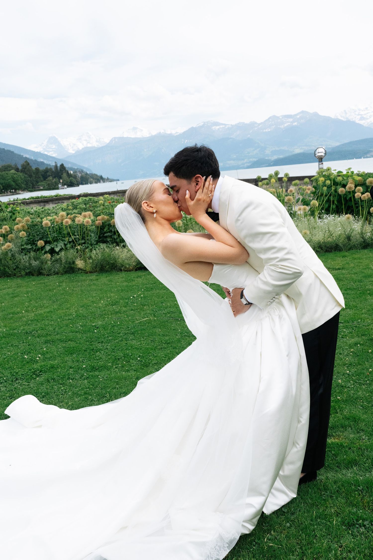 Bride and groom share a romantic dip kiss on a green lawn with a stunning alpine lake and mountain backdrop.