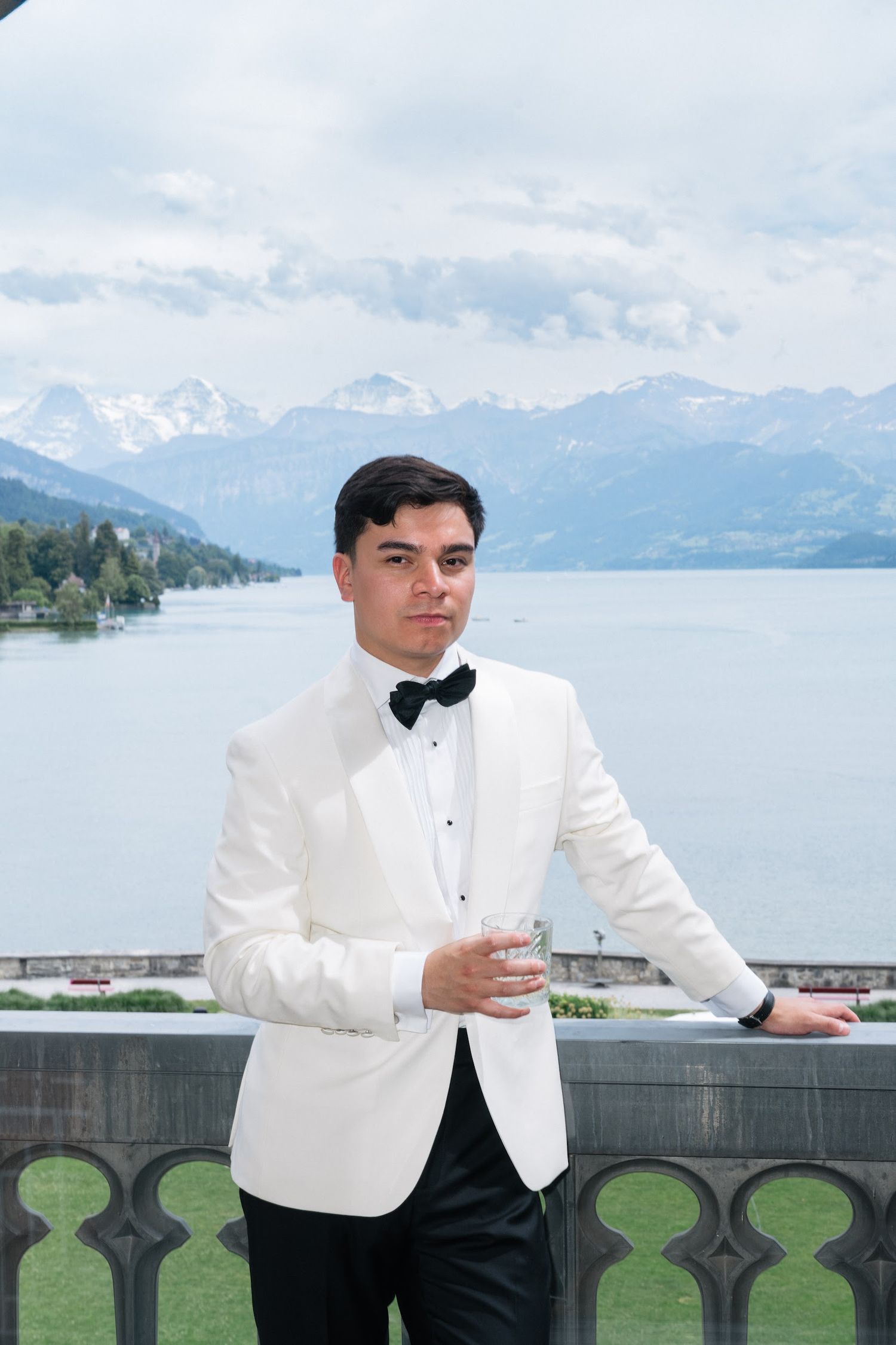 Dapper groom in white tuxedo jacket holding a drink on a balcony with alpine lake views.