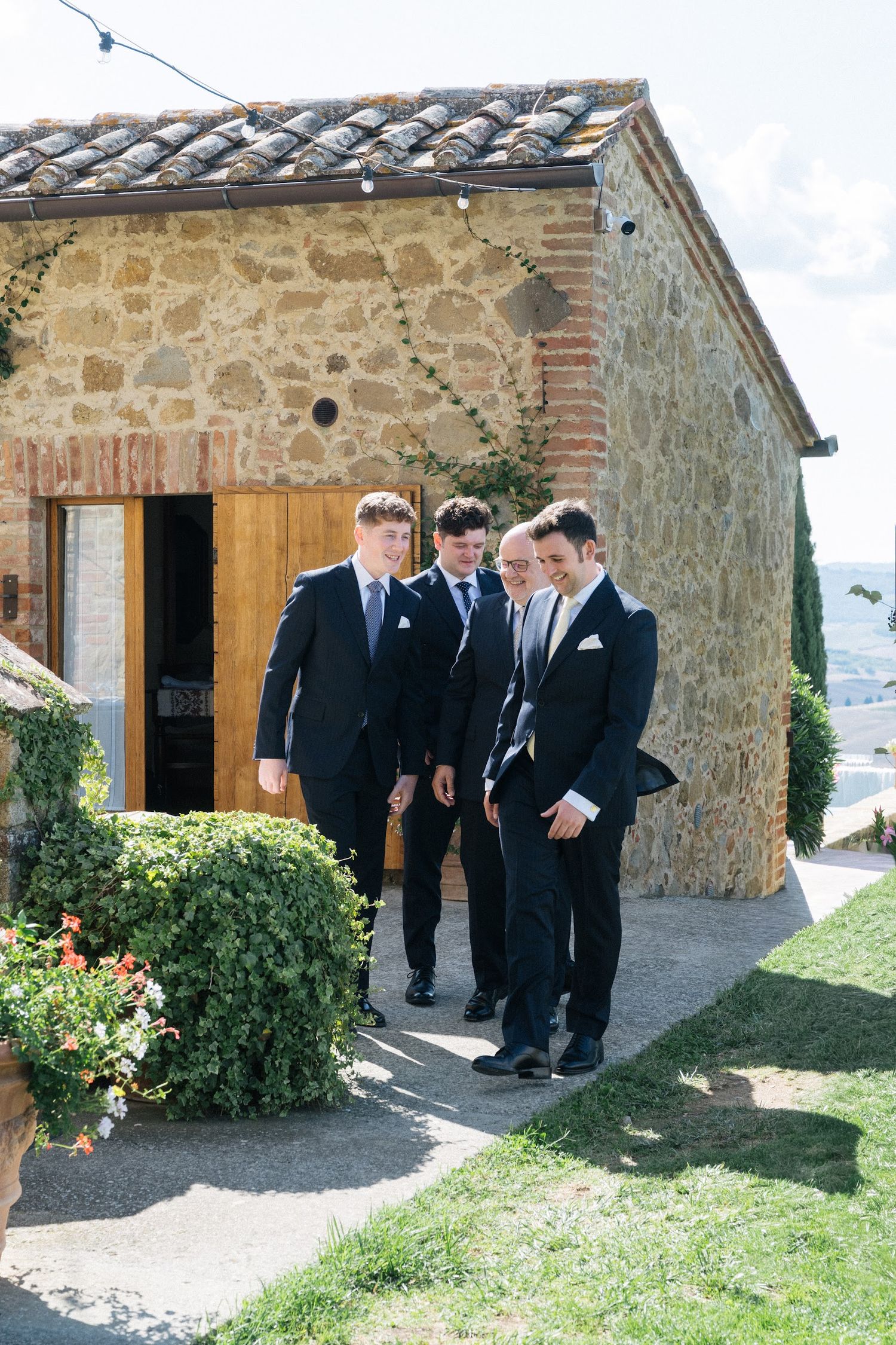 Groom and groomsmen in dark suits walking outside a rustic stone building on a sunny Italian wedding day.