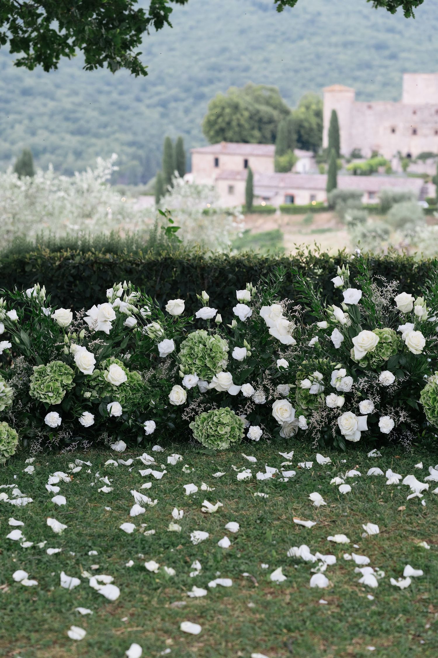 White roses and hydrangeas arranged along a hedge altar with a Tuscan castle in the background.