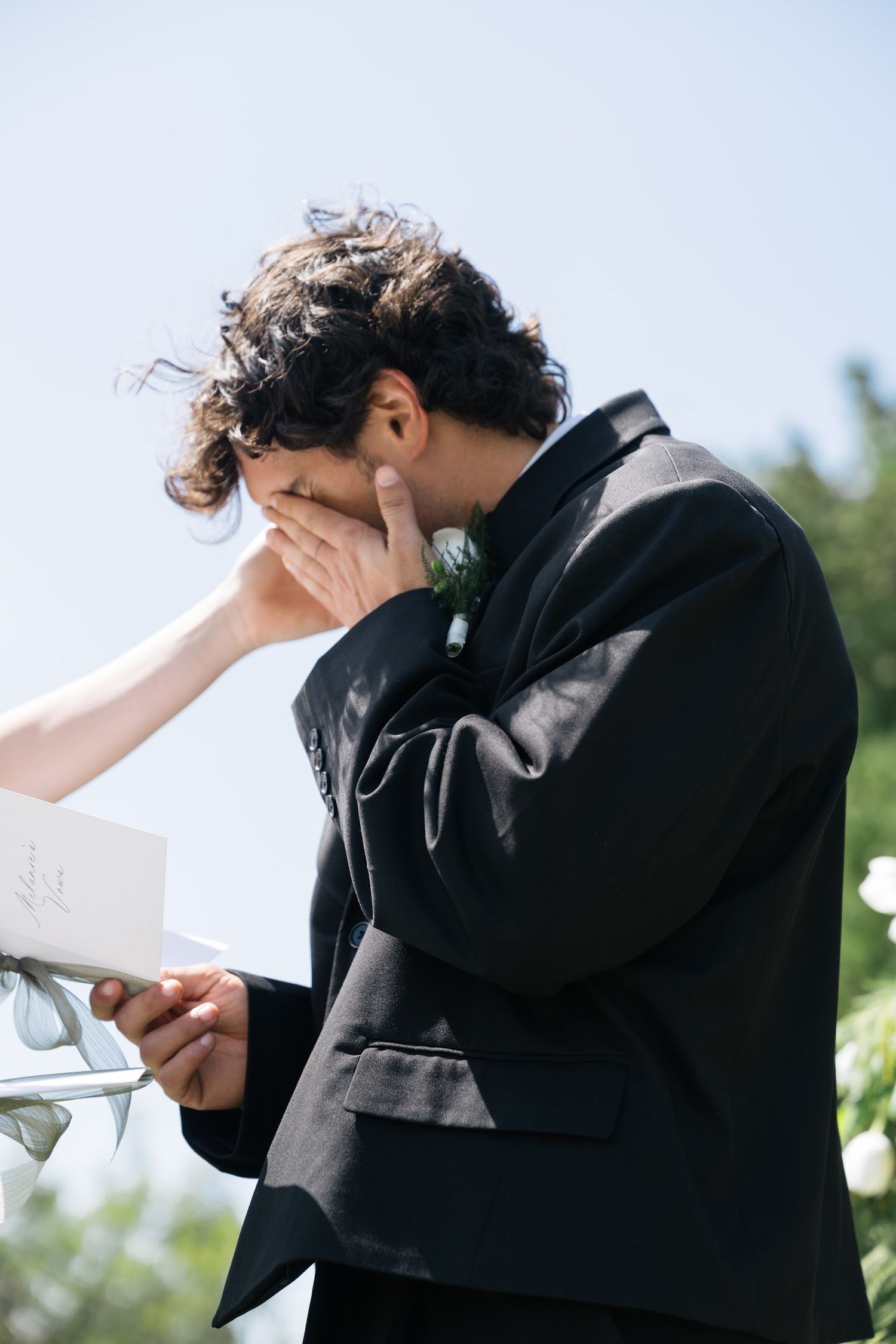 Emotional groom wipes tears while reading wedding vows at outdoor ceremony under bright blue sky.