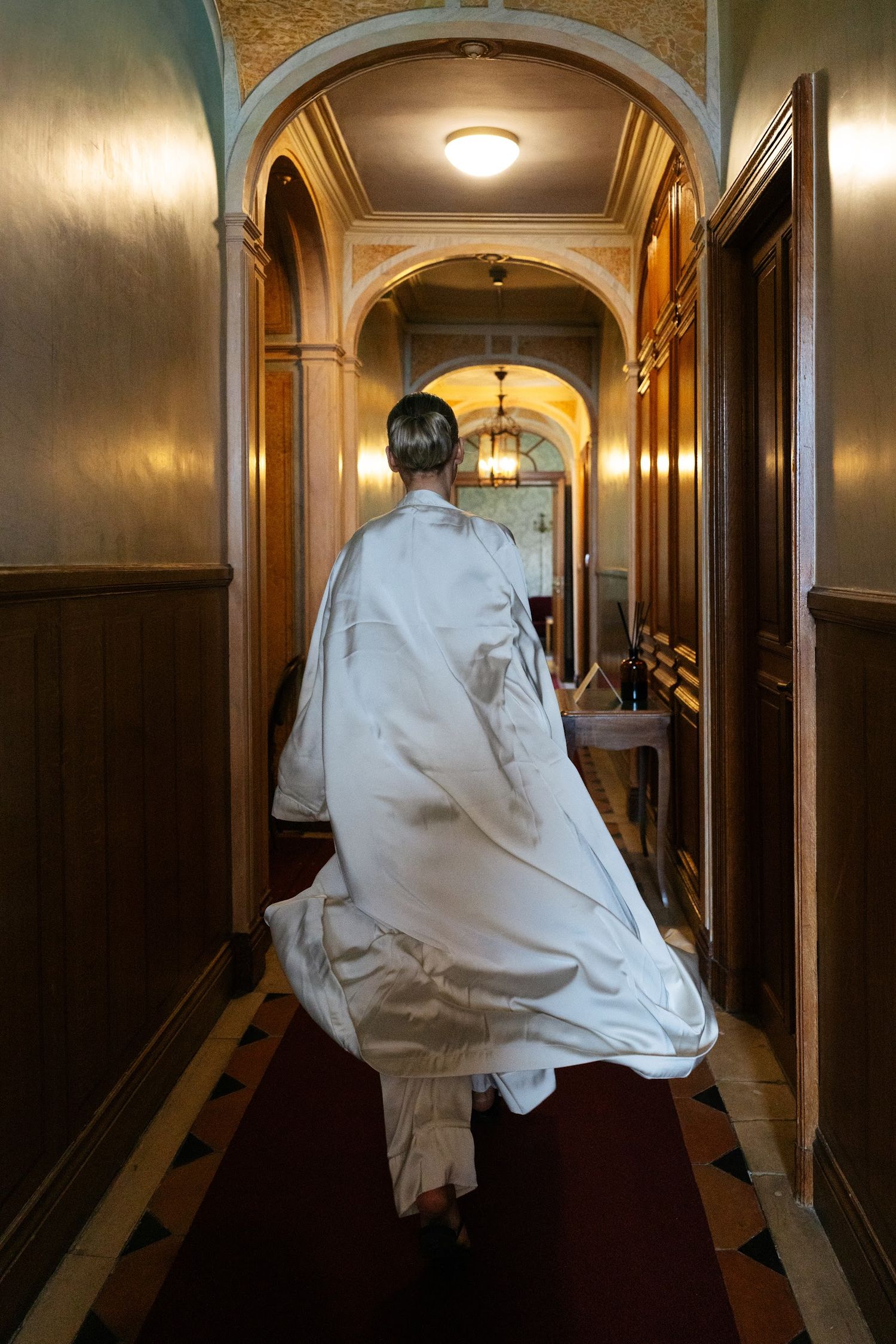 Bride in a flowing white robe walking down an ornate wood-paneled hotel corridor before her wedding.