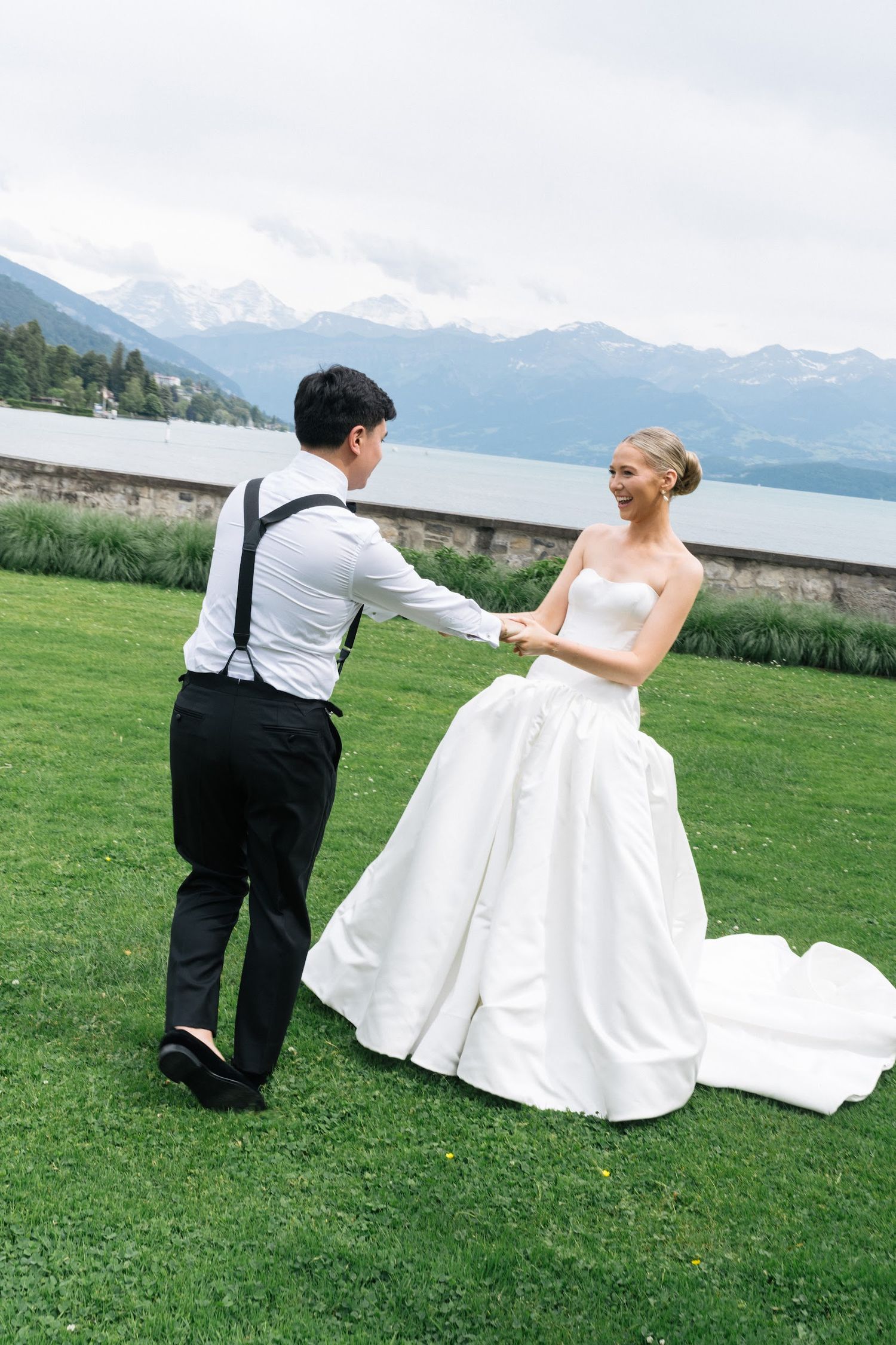 Groom in suspenders holding bride's hands on a green lawn with stunning alpine lake backdrop.