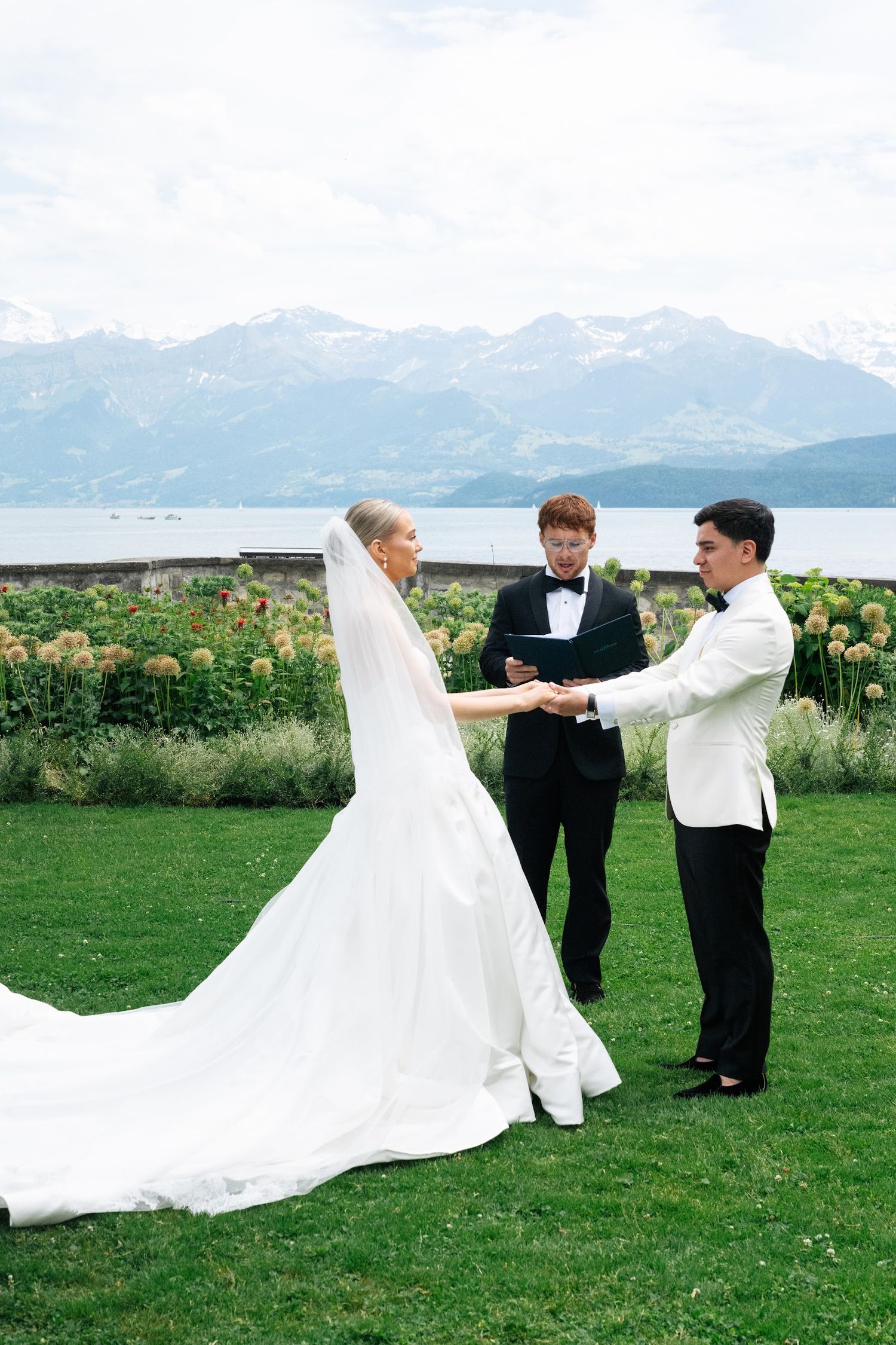 Bride and groom exchange vows and rings outdoors with an officiant, alpine lake and snow-capped mountains behind them.