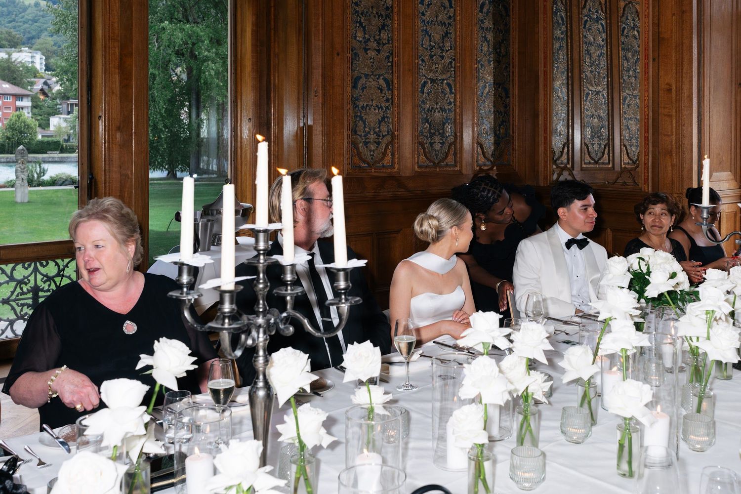 Wedding reception dinner at elegantly set table with white roses and candelabras in a wood-paneled room.