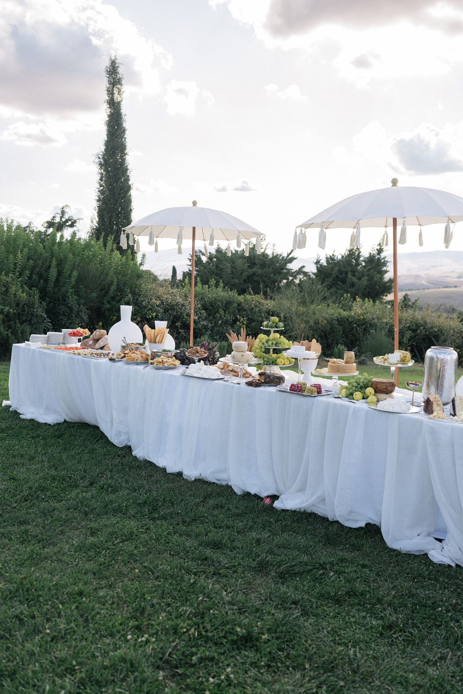 Outdoor wedding grazing tables with white draping and fringed umbrellas against rolling Tuscan countryside.