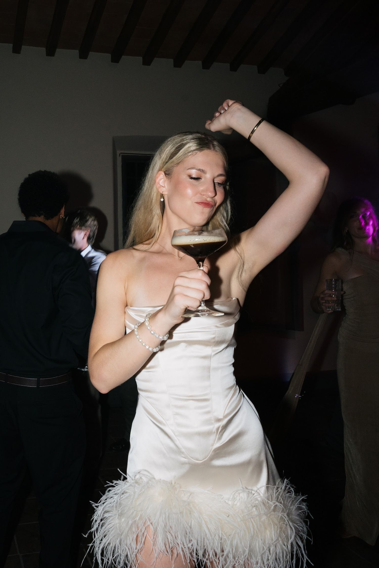 Blonde bride in satin feathered dress holds cocktail while dancing at wedding reception.