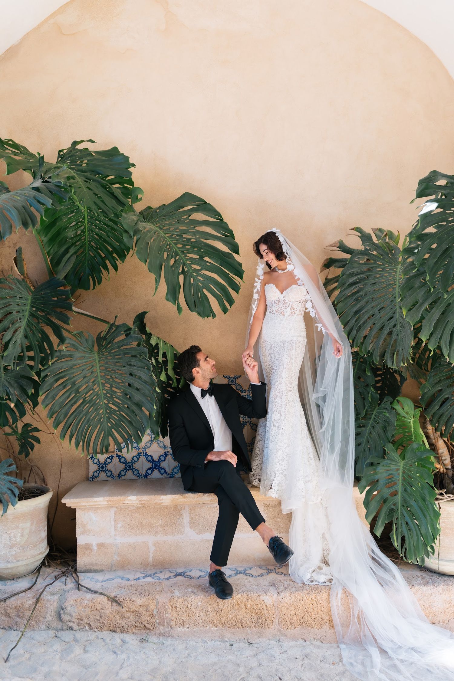 Bride in floral lace gown stands on steps with groom amid tropical monstera plants.
