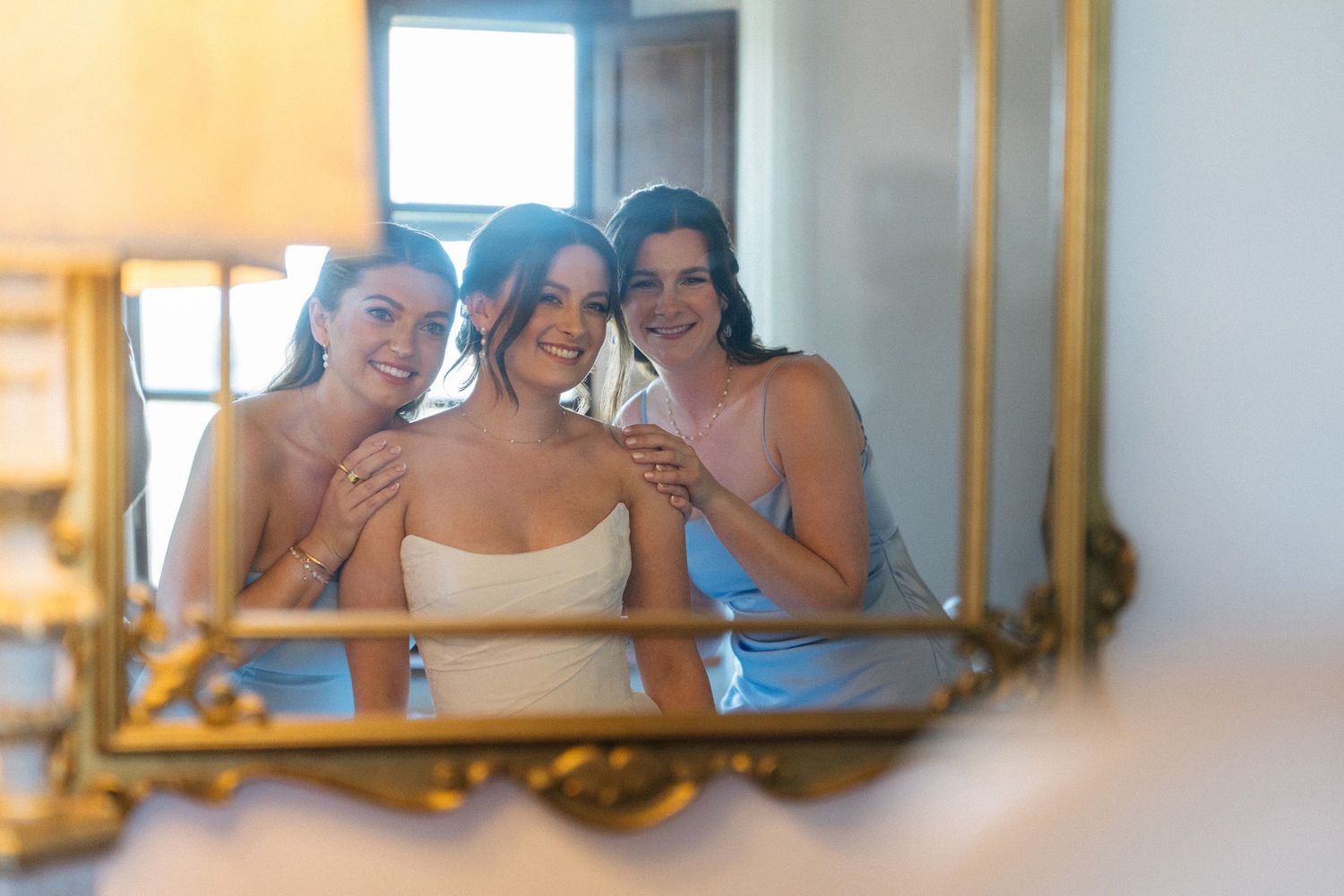 Bride and two bridesmaids in blue dresses smiling together reflected in ornate gold mirror.