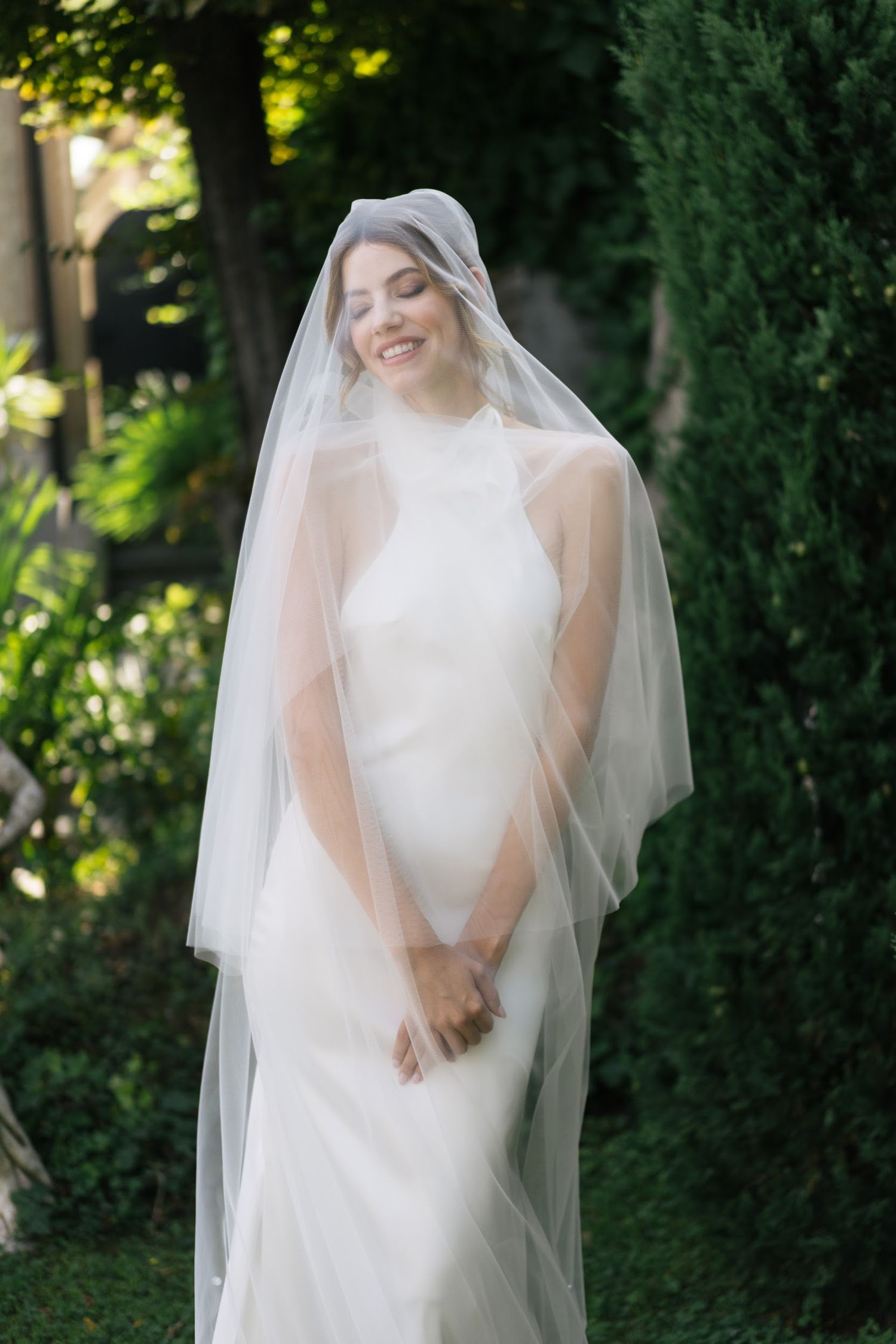 Smiling bride in sleek white gown and flowing tulle veil poses outdoors among lush green trees.
