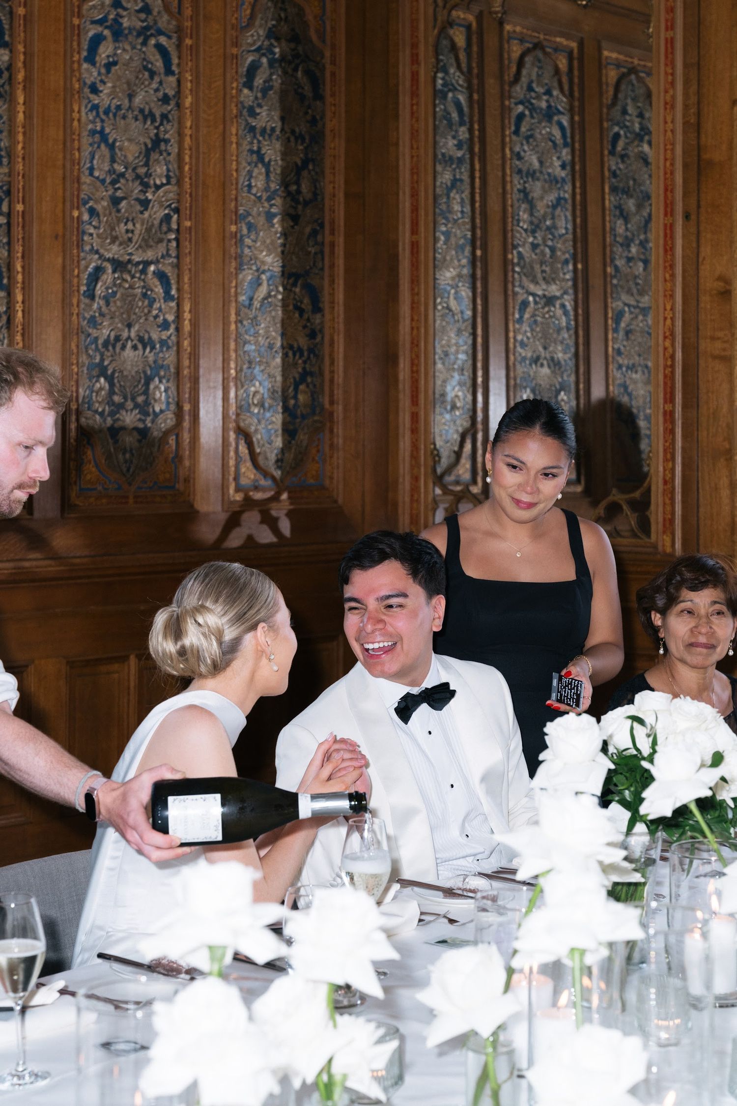 Groom in white tuxedo laughs with guests at elegant wedding reception with white floral centerpieces.