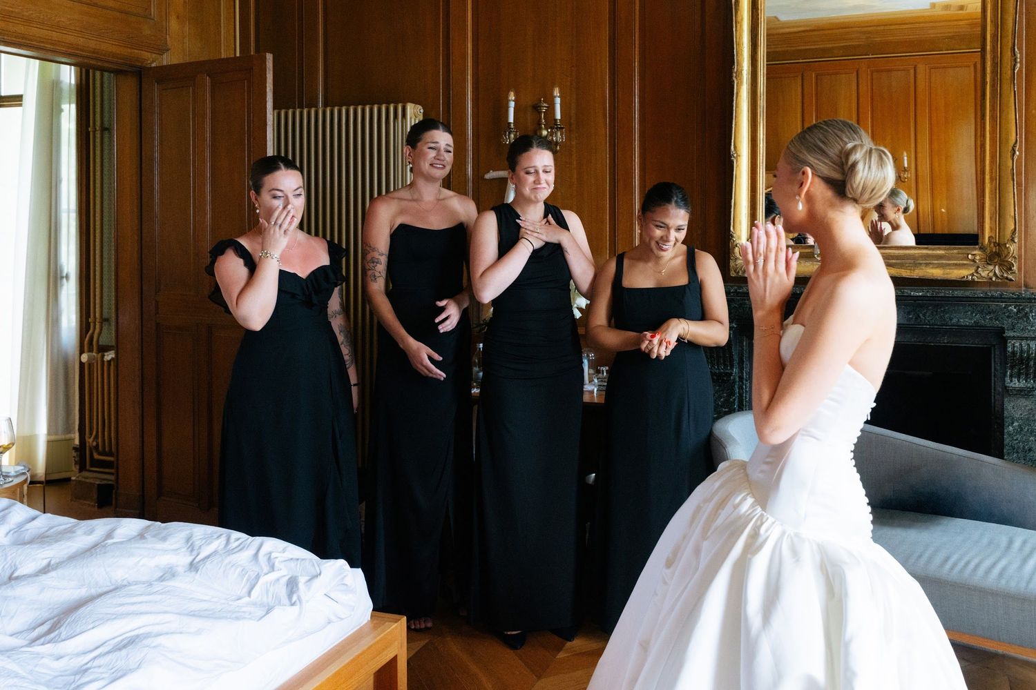 Emotional bridesmaids in black dresses reacting to bride's reveal in wood-paneled room.