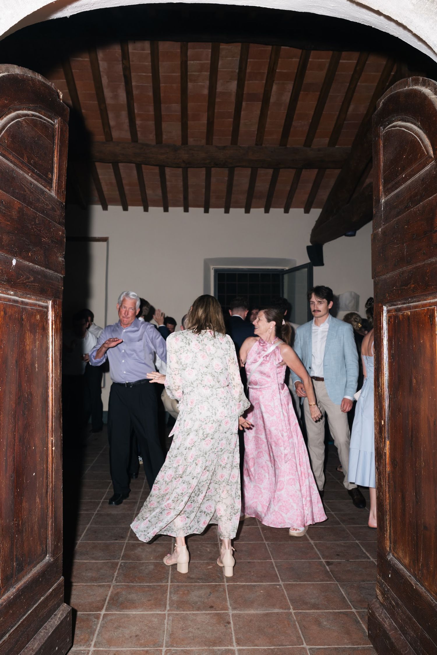 Wedding guests in formal attire dancing through ornate wooden doorway at rustic venue.