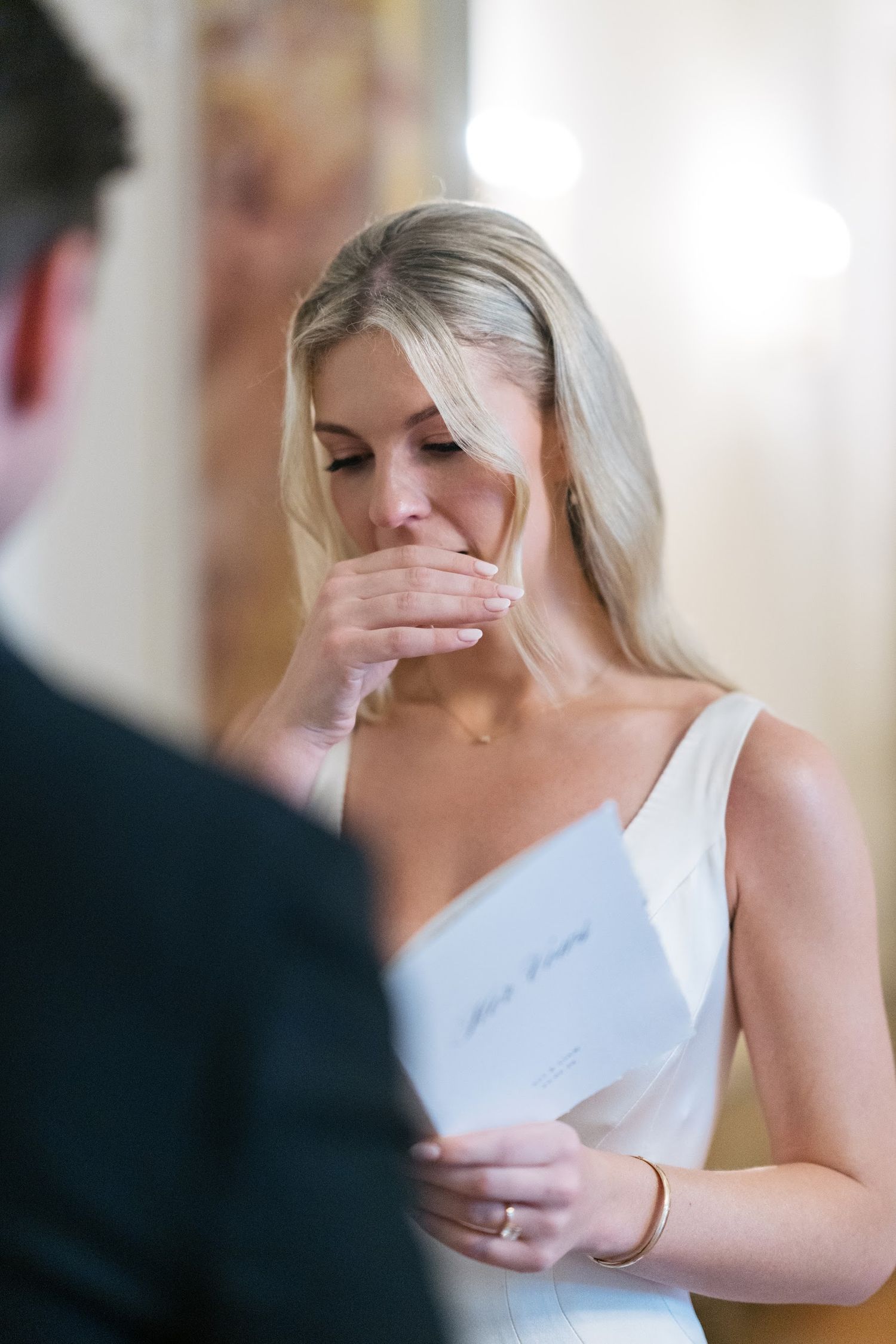 An emotional blonde bride covers her mouth while reading wedding vows from a card during ceremony.