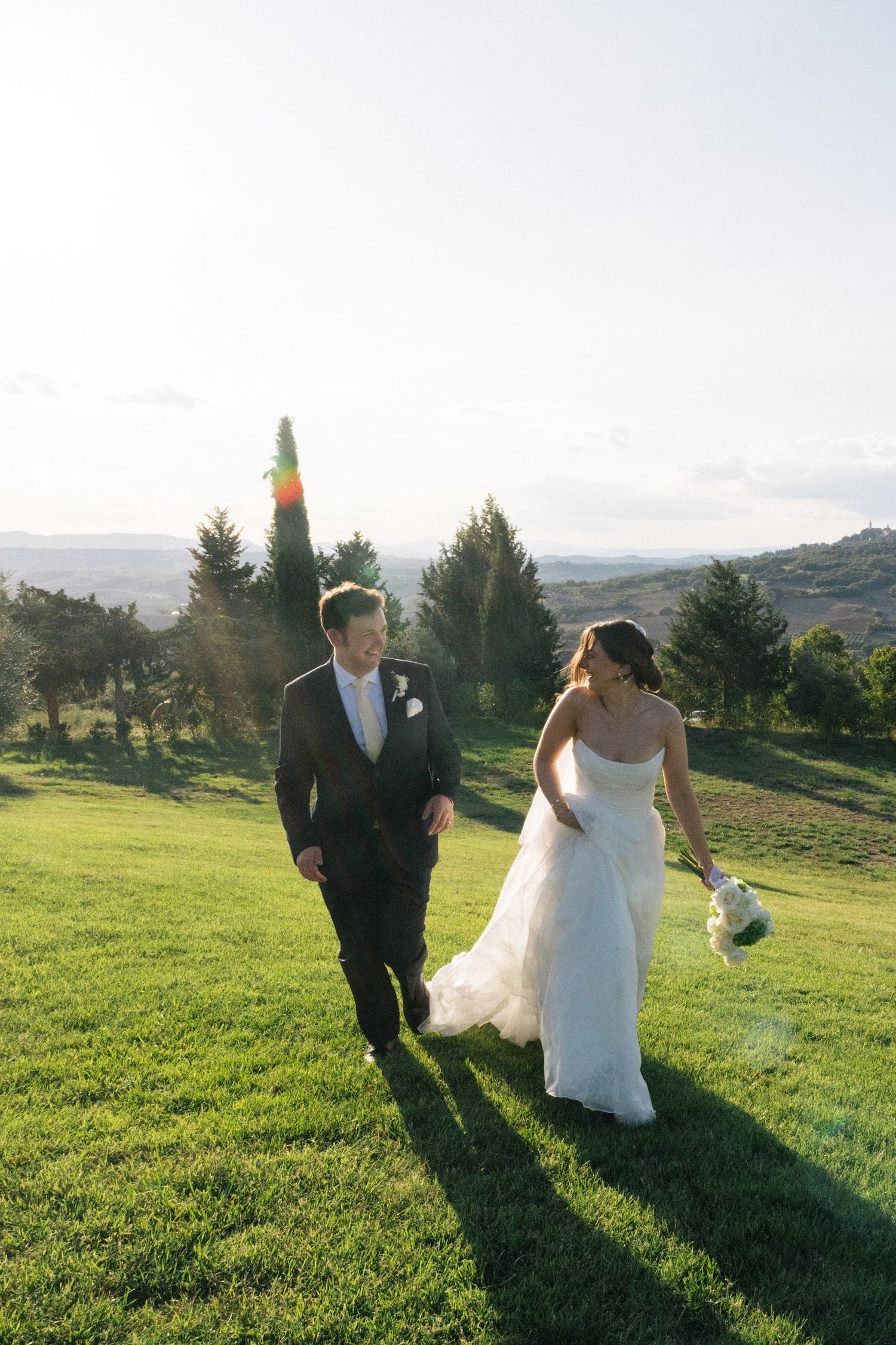 Bride and groom walk joyfully across sunlit green hillside with panoramic Tuscan landscape at sunset.