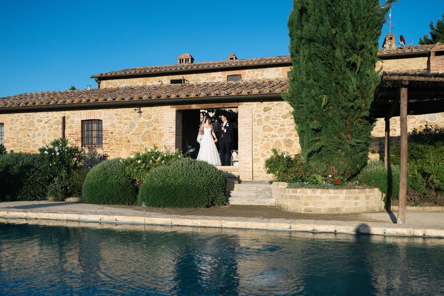 Newlyweds exit rustic stone Tuscan villa beside a serene swimming pool on a clear sunny day.