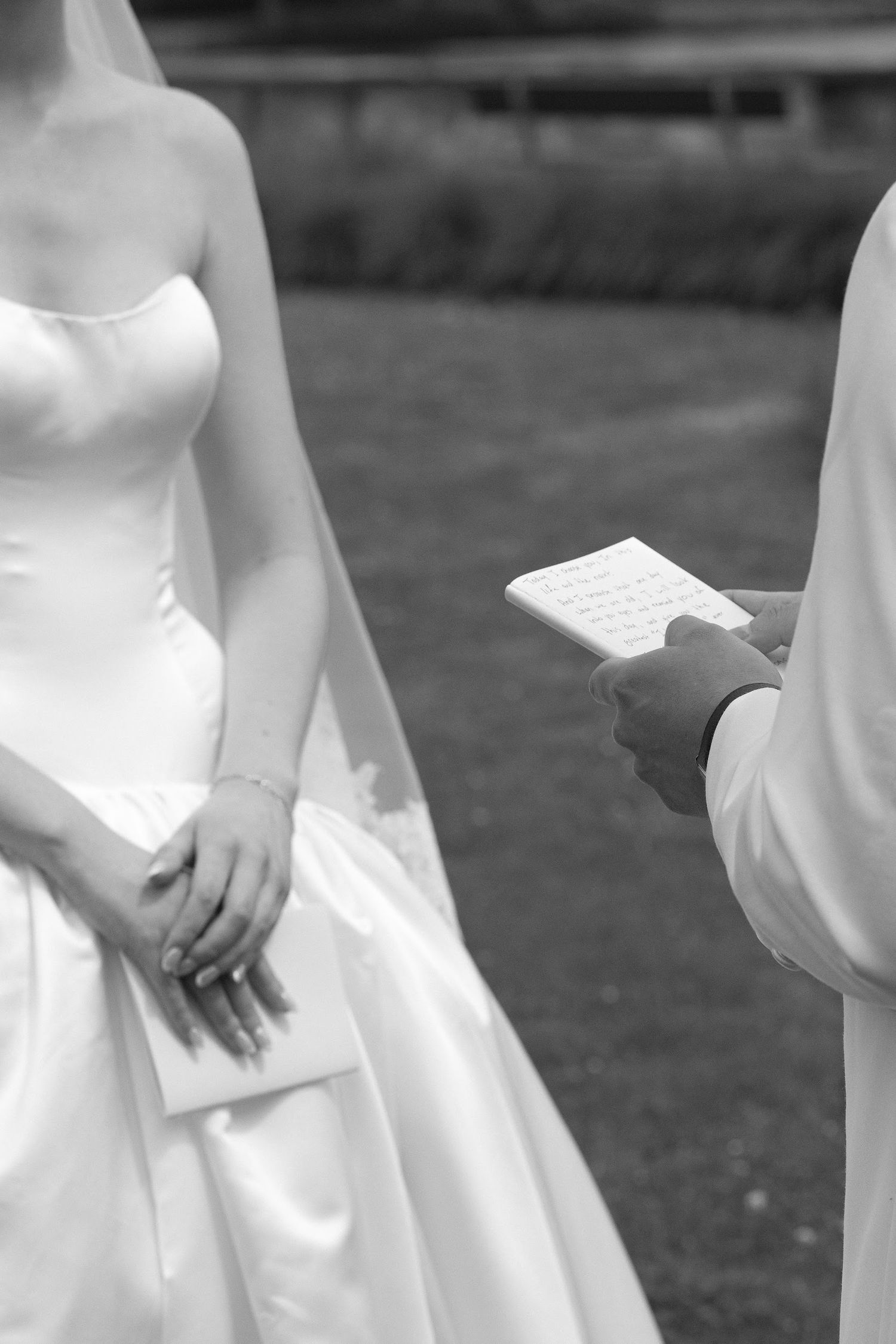 Black and white close-up of groom reading vows from card while bride in strapless white gown listens during ceremony.