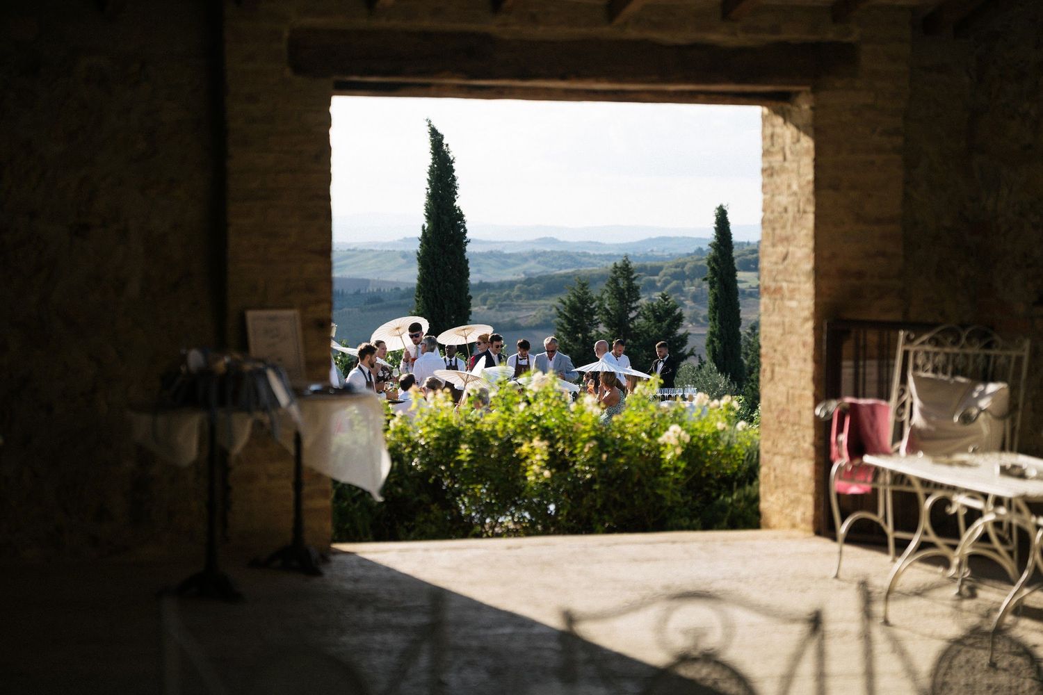 Wedding guests gathered in sunlit Tuscan courtyard framed by stone archway with scenic valley view.