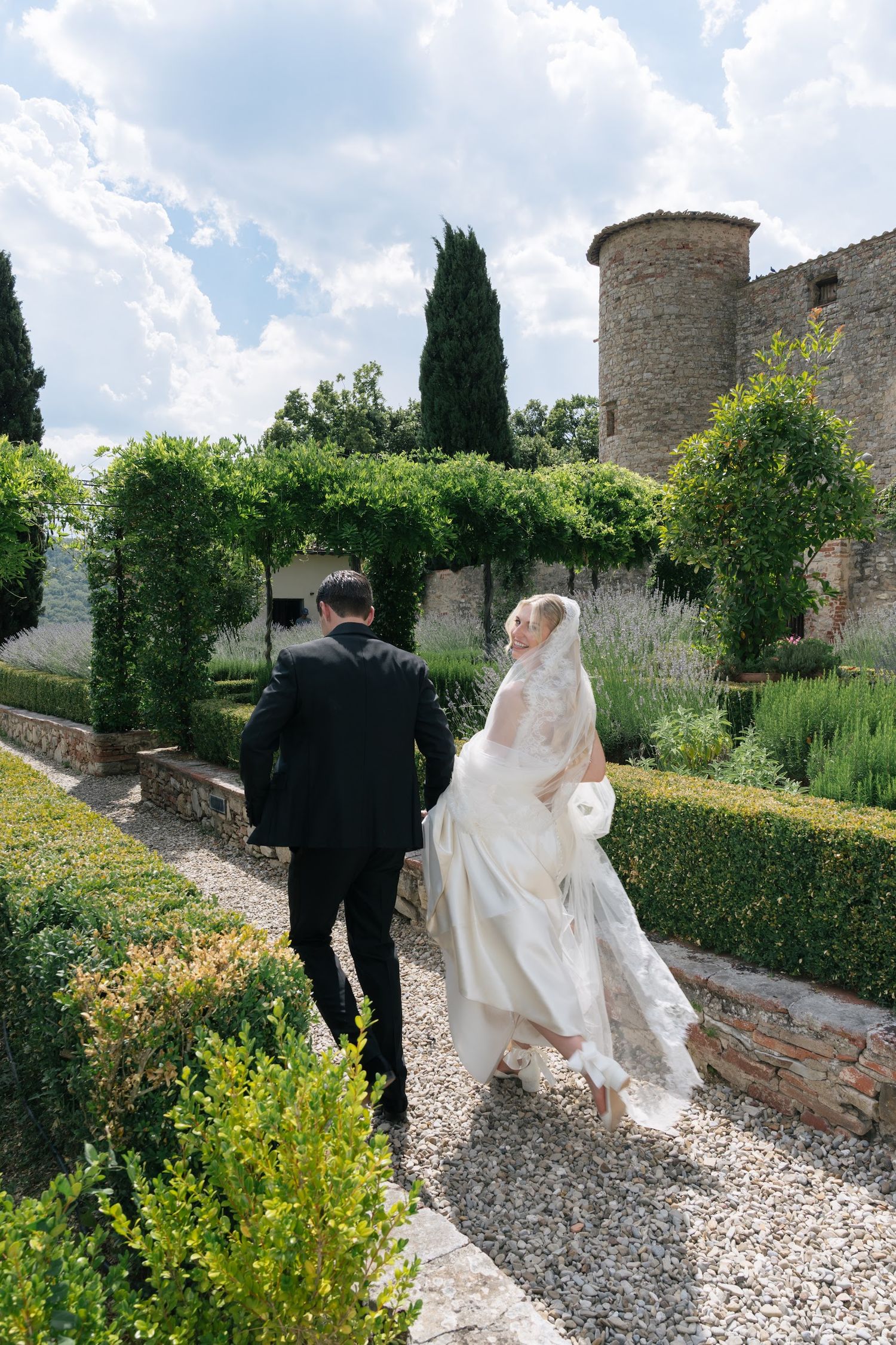 Bride in a flowing white gown and groom in a tuxedo walk through a Tuscan castle garden.