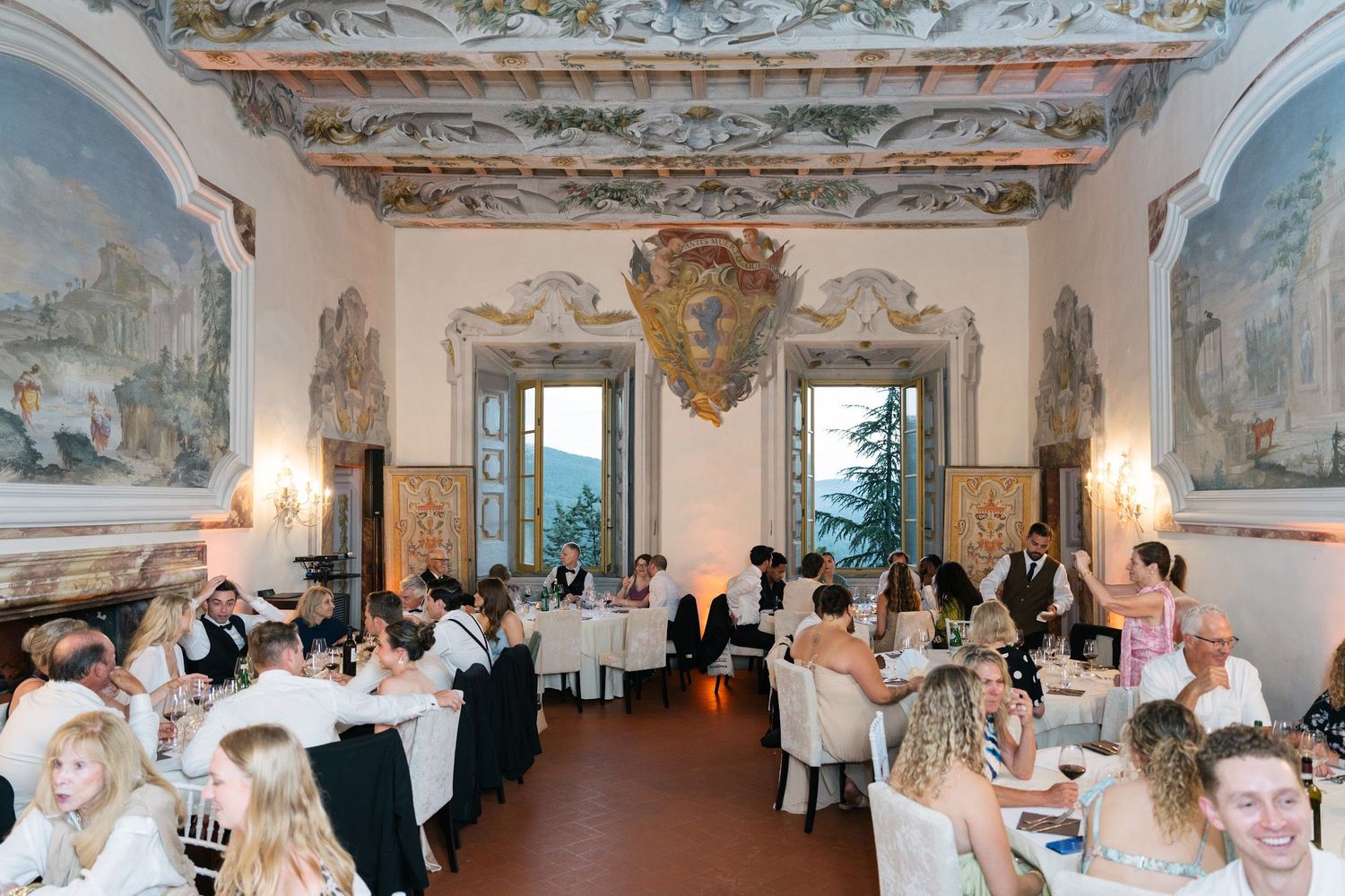 Guests dining in ornate frescoed ballroom at luxurious Italian villa wedding reception.